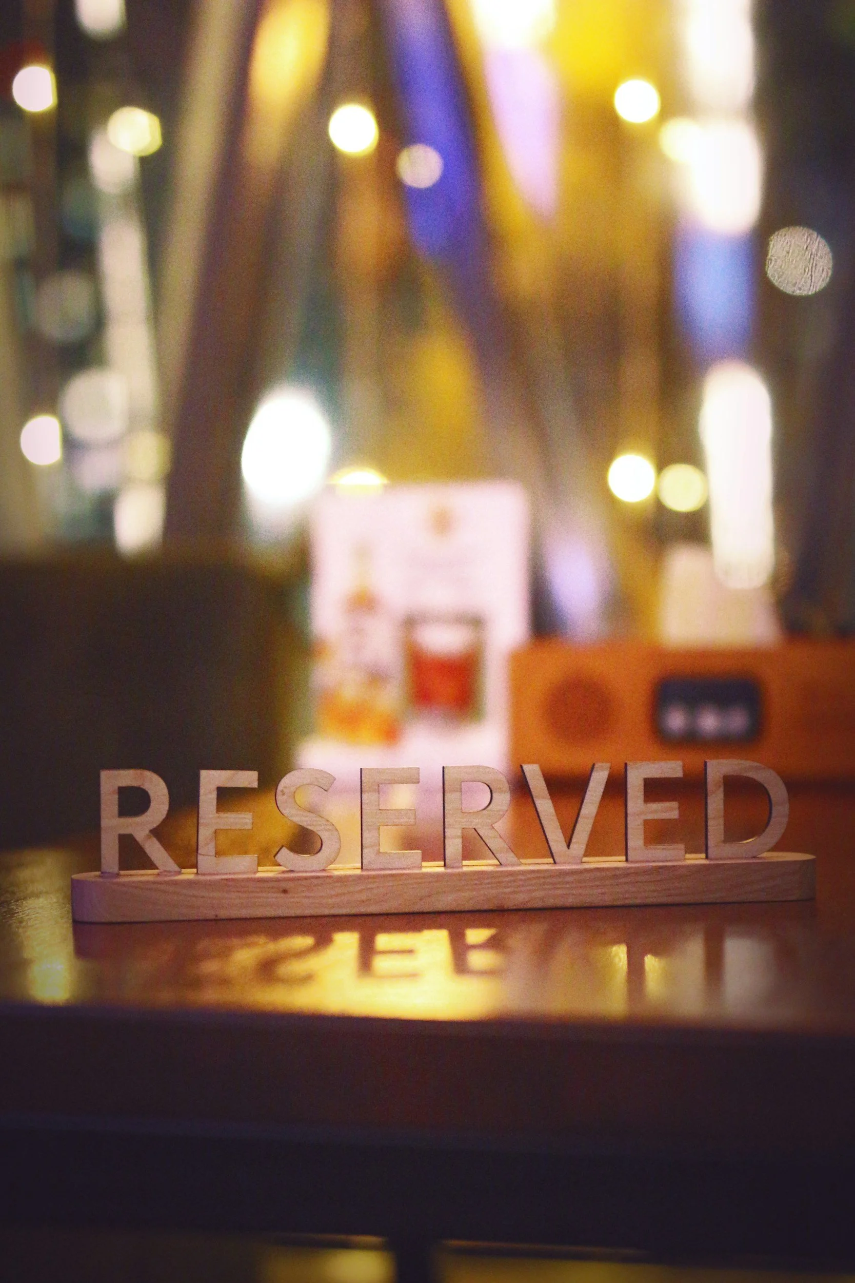A wooden 'RESERVED' sign on a table in a dimly lit restaurant with blurred colorful lights and menus in the background.