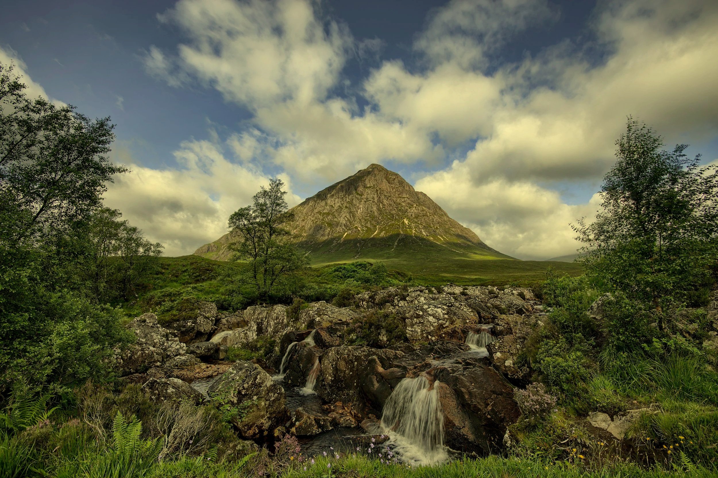 A mountain surrounded by greenery with a stream flowing over rocks in the foreground and a partly cloudy sky above.