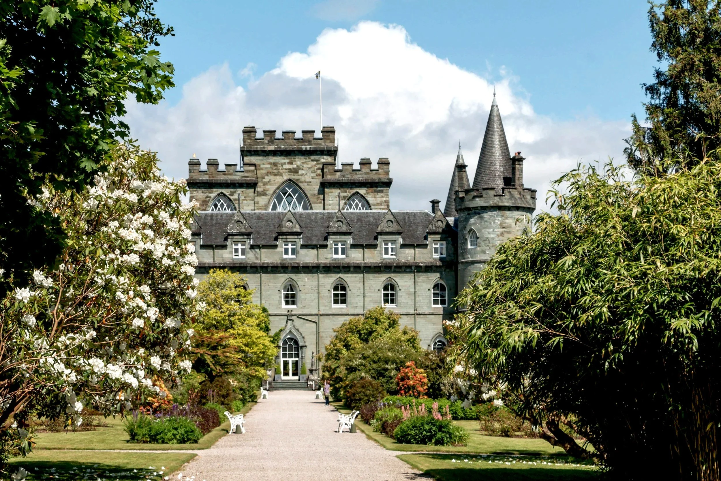 A large, historic castle with stone walls, towers, and pointed turrets, surrounded by a garden with trees and flowering bushes, under a partly cloudy sky.