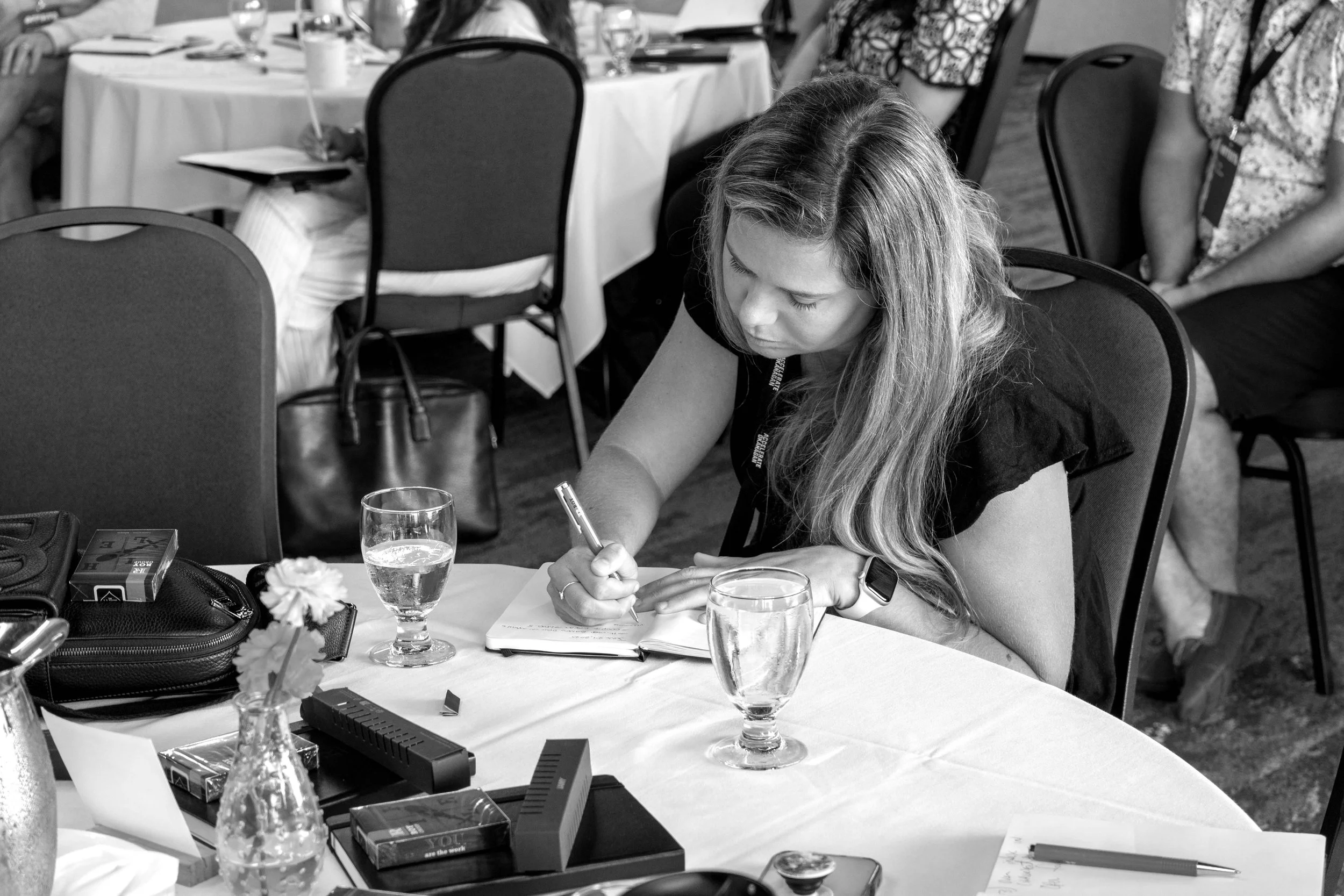 A woman with long hair writing in a notebook at a round table during a meeting or conference, with glasses of water and office supplies on the table.
