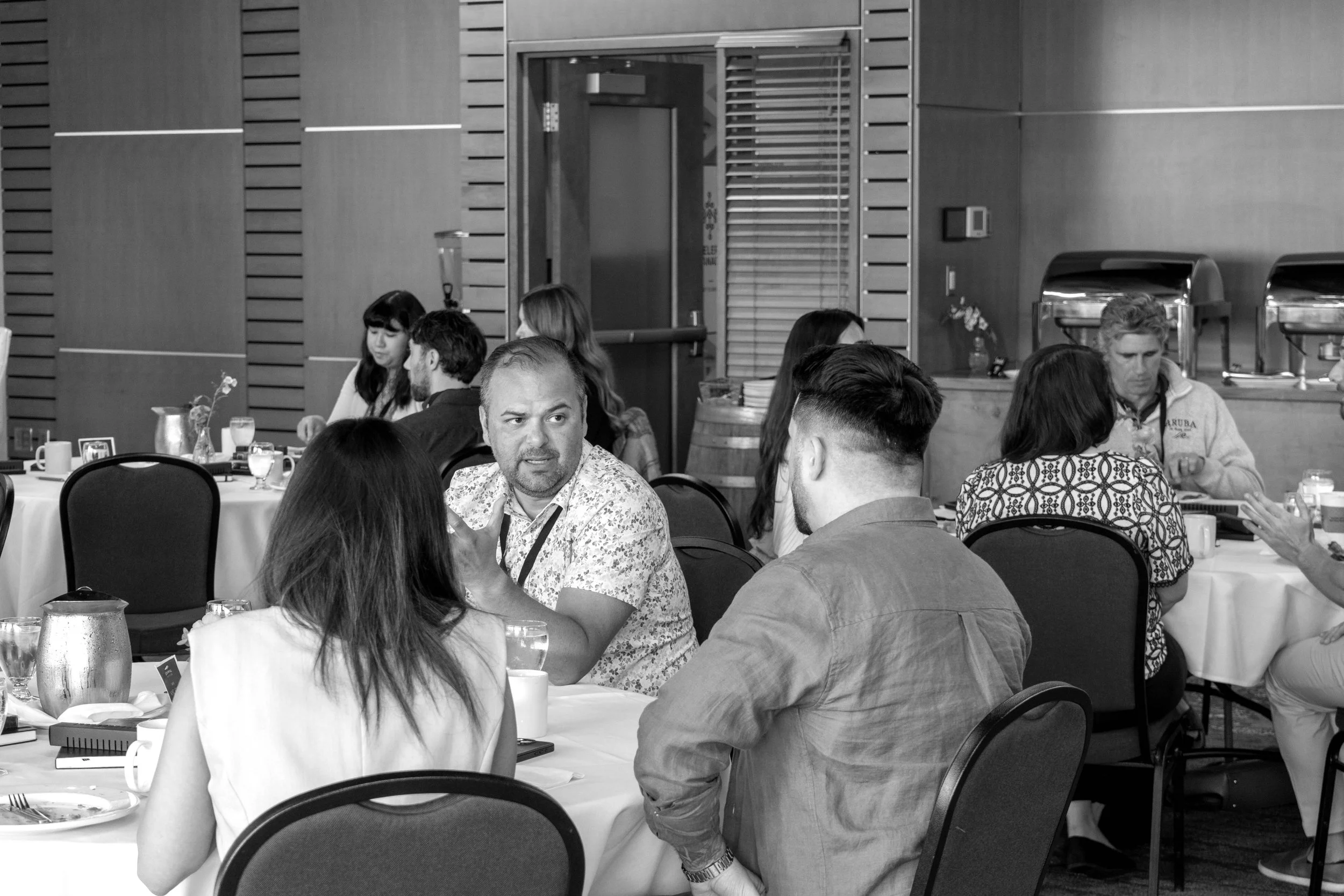 People sitting at round tables in a restaurant or banquet hall, engaged in conversation, with some eating and drinking, and a buffet station in the background.