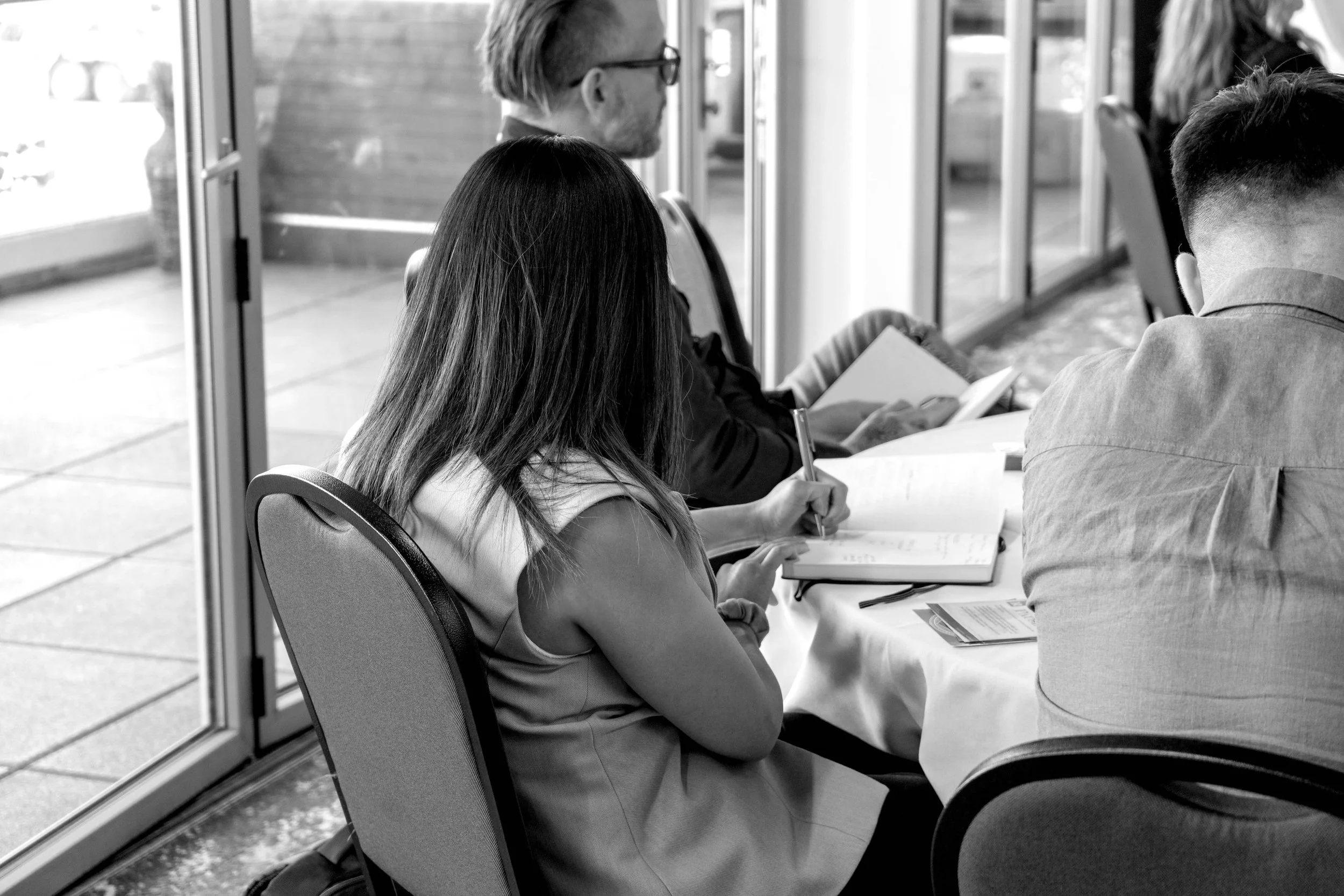 A group of people sitting around a table, taking notes and using laptops in a bright conference room with large windows.