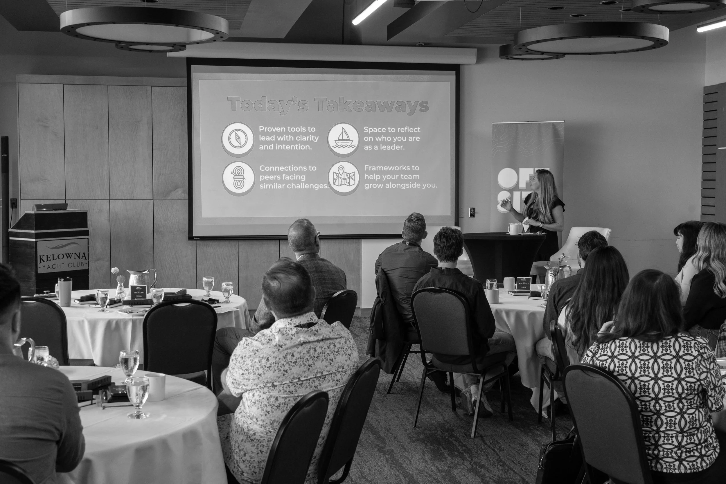 A woman presenting at a conference with slides titled 'Today's Takeaways' on a large screen. The audience is seated at round tables with drinks and notepads.