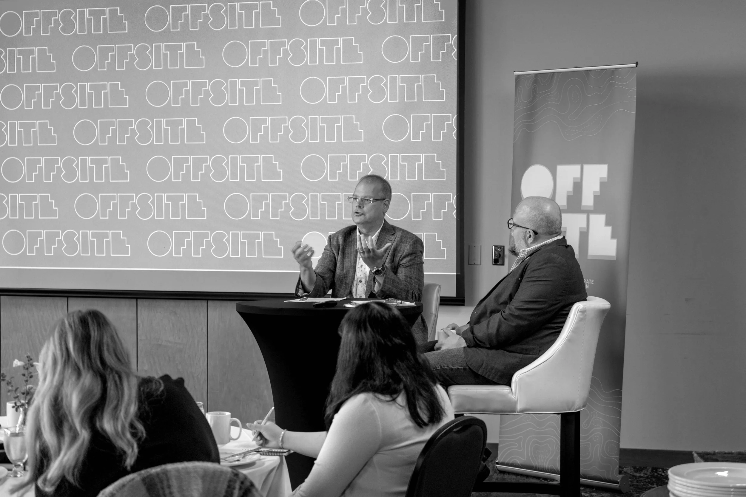 Two men speaking at a panel discussion during a conference, with a presentation screen behind them displaying the OFFSITE logo. Attendees are seated at tables, taking notes.