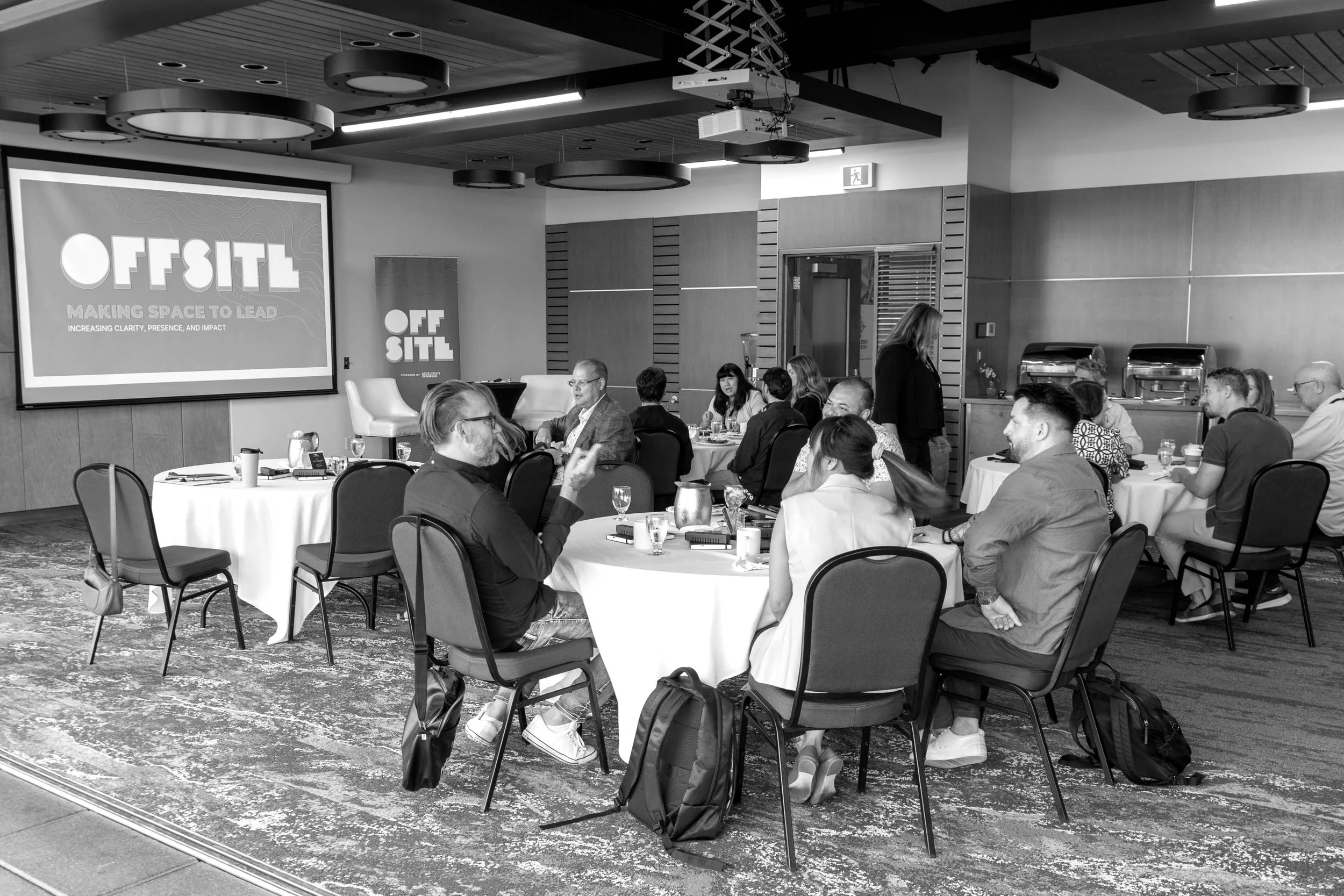 A conference room with people sitting around round tables during a presentation. A large screen displays the 'OFFSITE' logo and the phrase 'Making Space to Lead.' Attendees are engaged in conversation, some with notebooks and drinks, with a woman sta