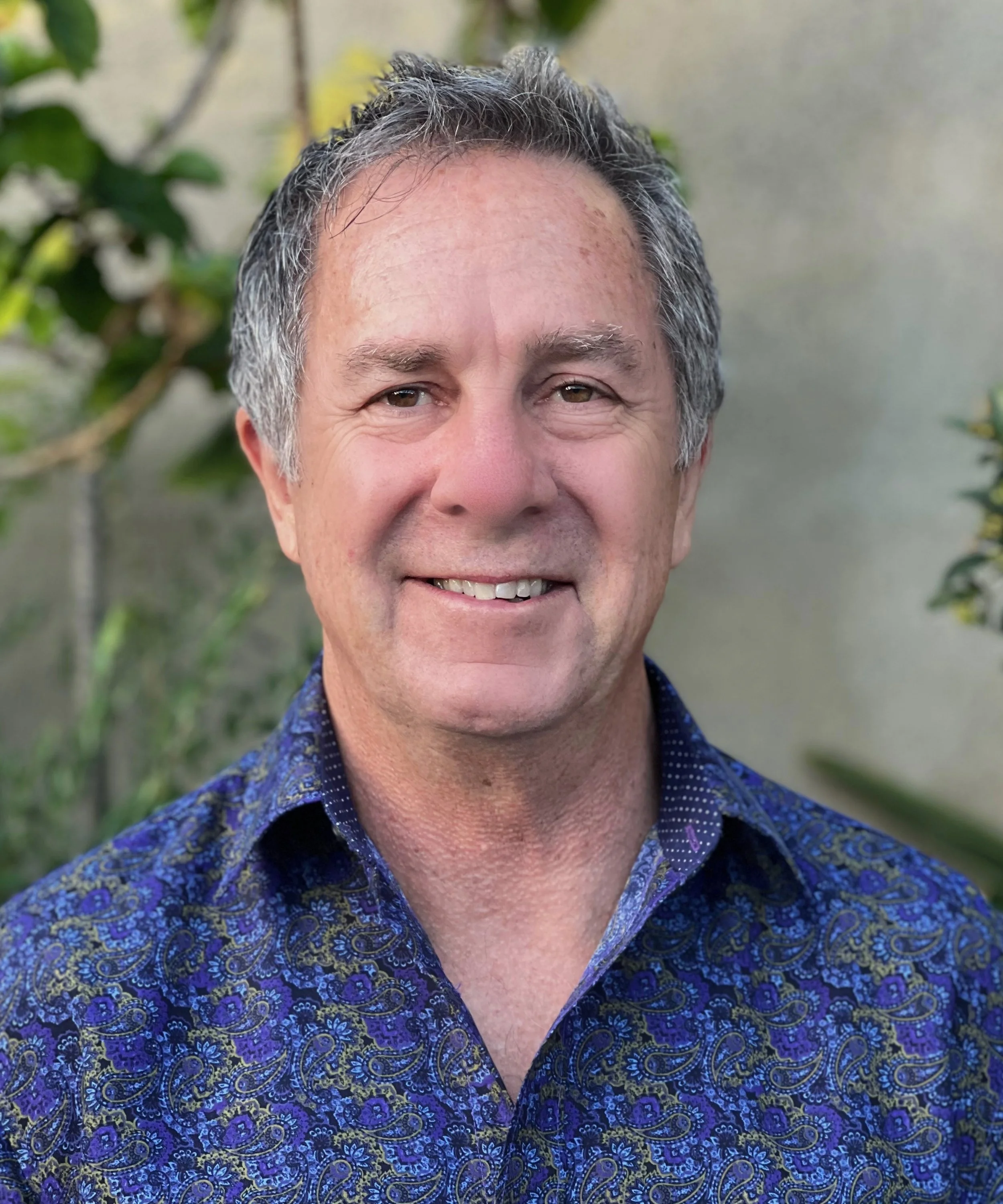 A middle-aged man with gray hair, smiling, wearing a blue patterned shirt, outdoors with greenery in the background.