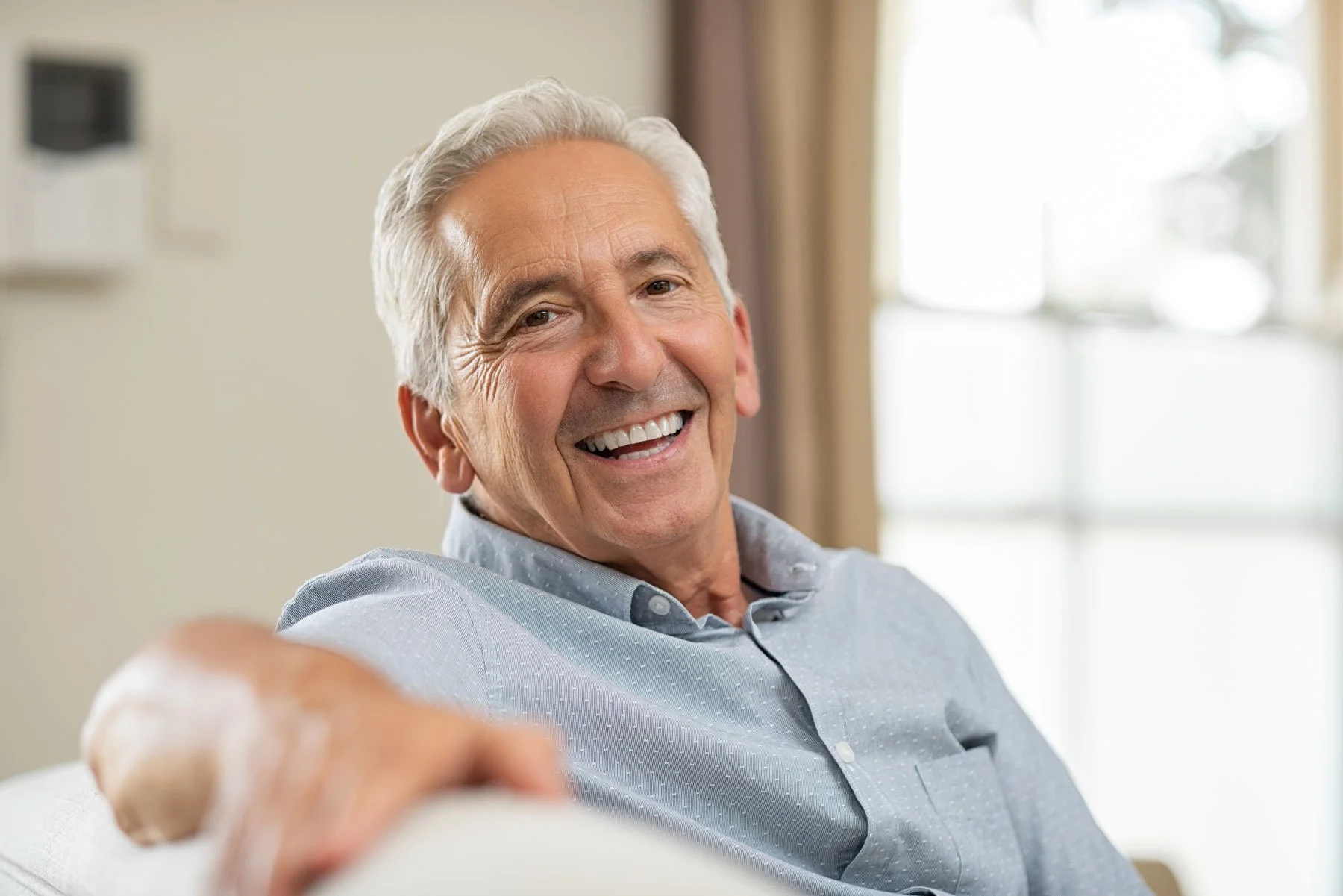 An older man with gray hair and a broad smile, wearing a light blue button-up shirt, sitting and reaching out with his right hand in a bright room with large windows and curtains.