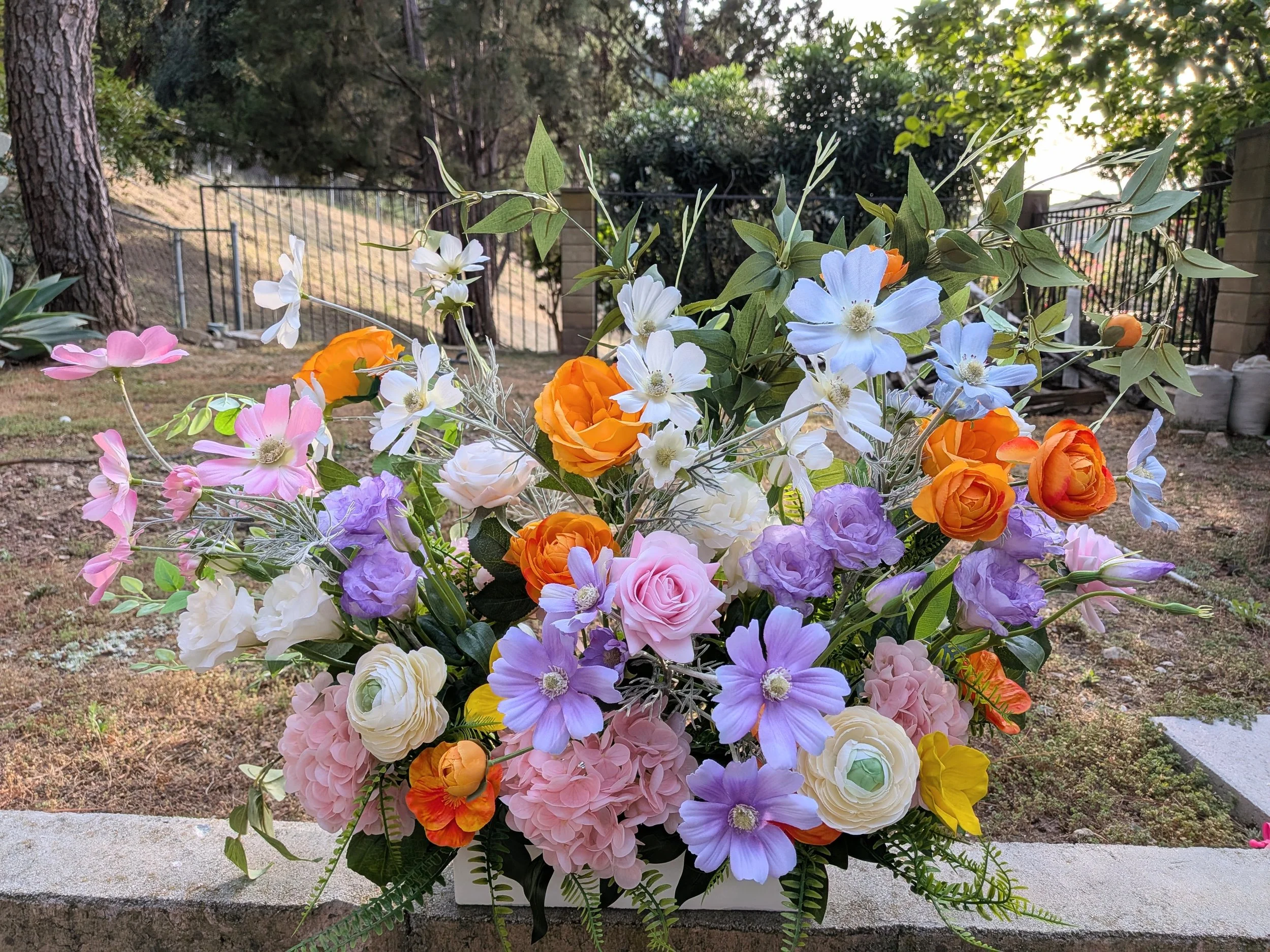 Colorful bouquet of various flowers, including orange roses, pink, purple, and white blooms, arranged outdoors on a stone ledge with trees and a fence in the background.