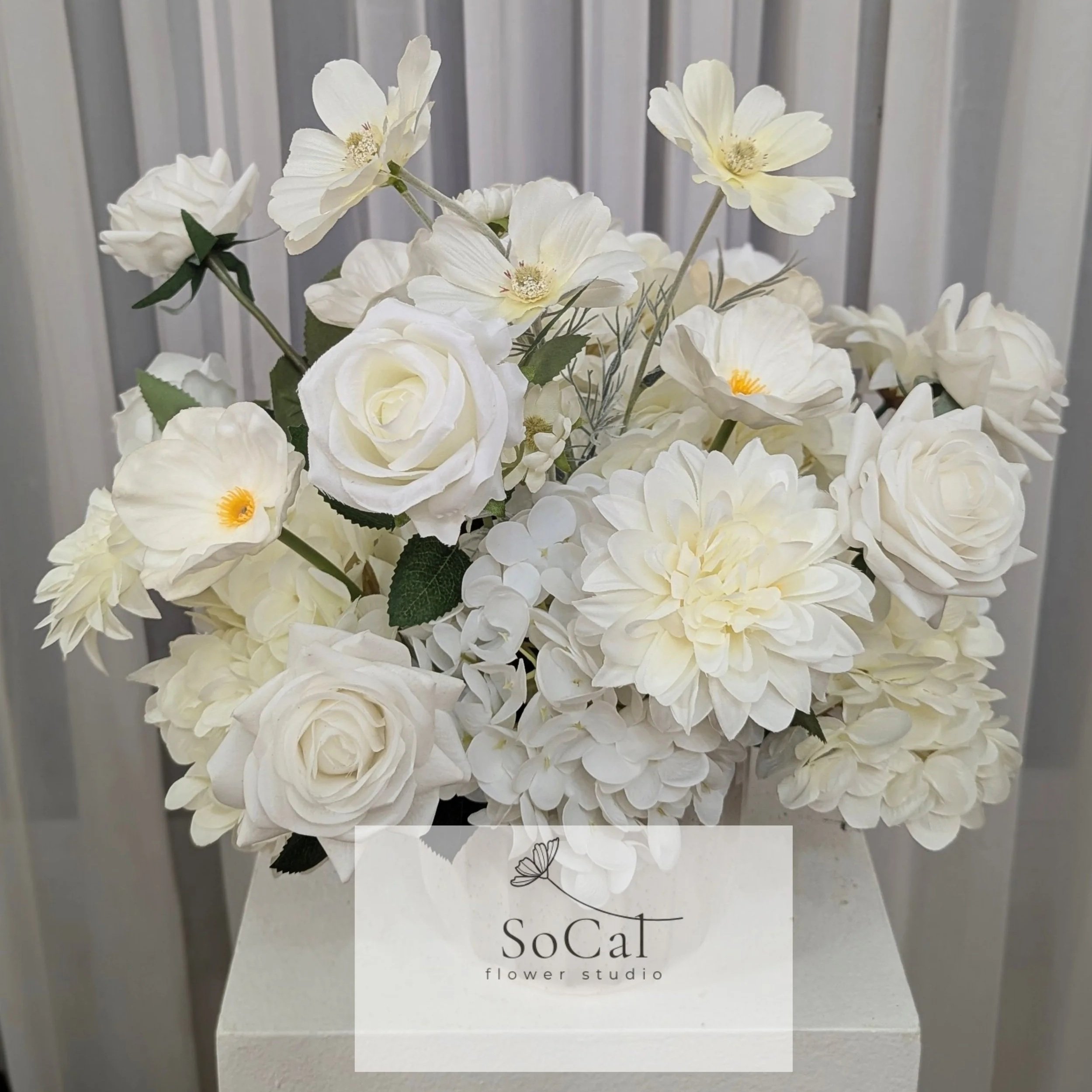 A bouquet of white flowers including roses, poppies, dahlias, and other blooms in a white vase with a SoCal flower studio card, set against a light gray curtain background.