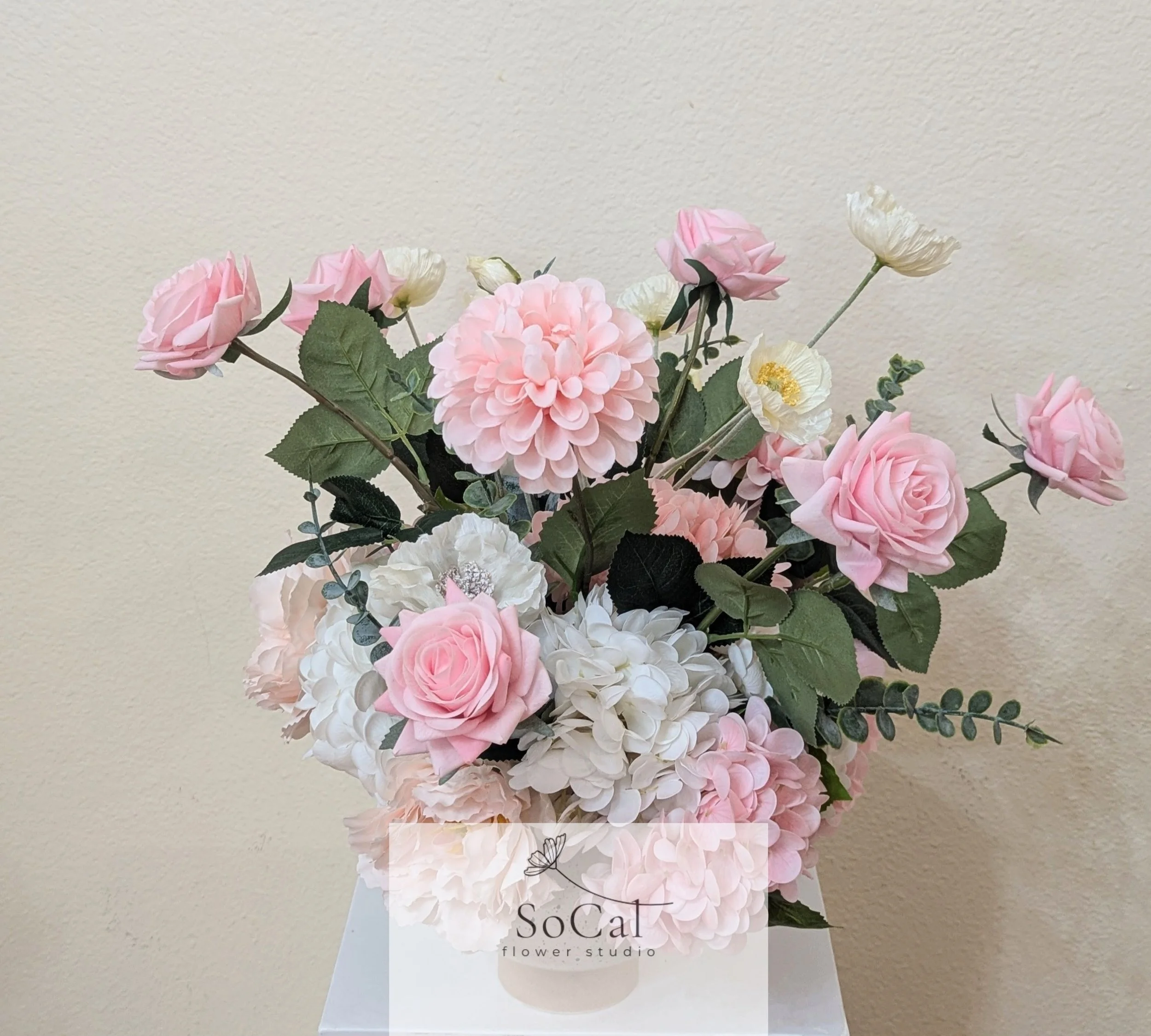 A bouquet of pink and white roses and hydrangeas in a white box with a 'SoCal flower studio' label, against a beige wall.