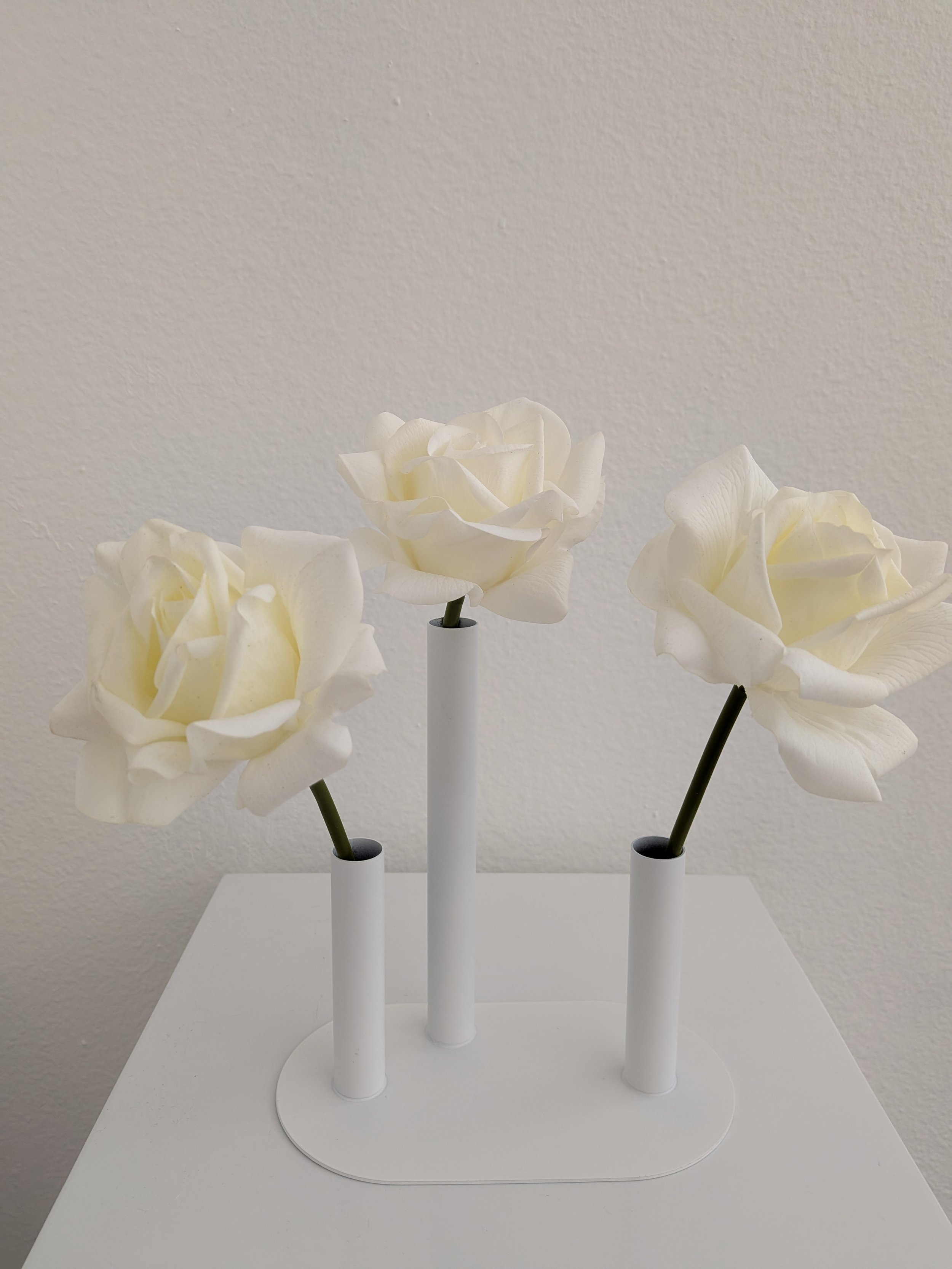 Three white artificial roses on black stems in white cylindrical vases on a white table against a beige wall.