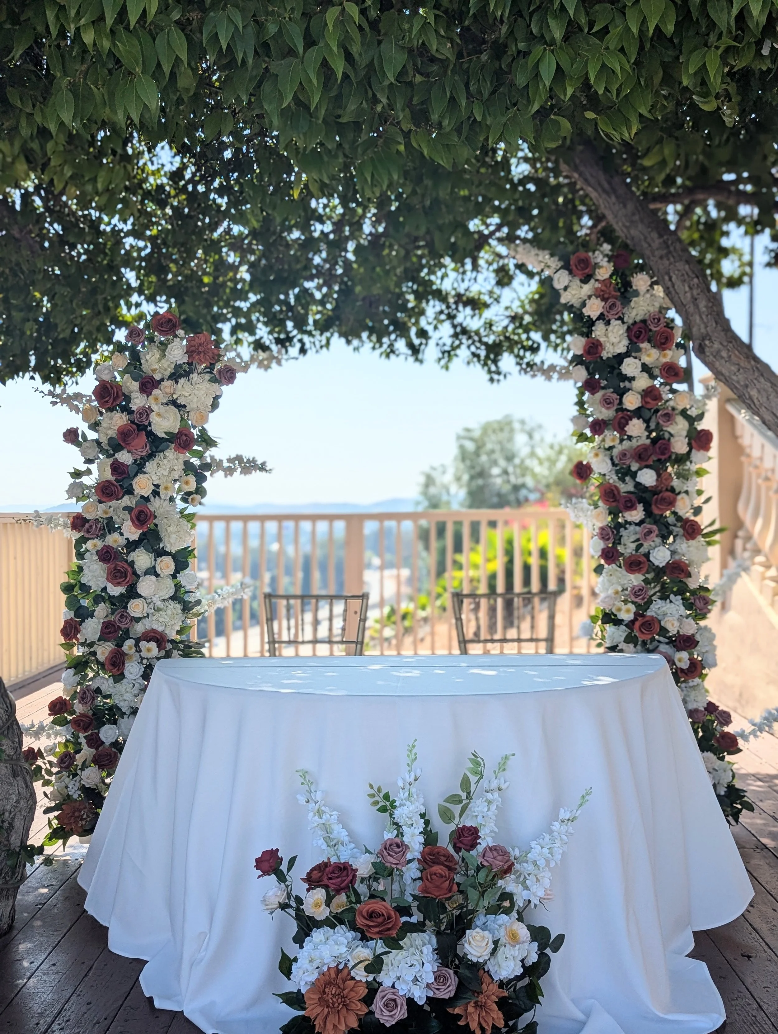 Decorated wedding altar with floral arch, table, and chairs on a wooden deck with a scenic view.