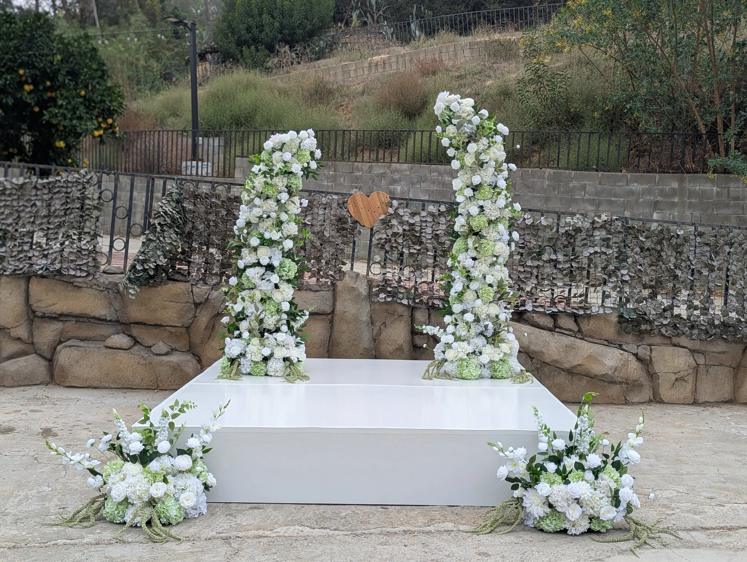 Outdoor wedding altar decorated with white flowers and greenery, featuring two flower arrangements at the base and floral arches behind, with a wooden heart ornament hanging in the center.