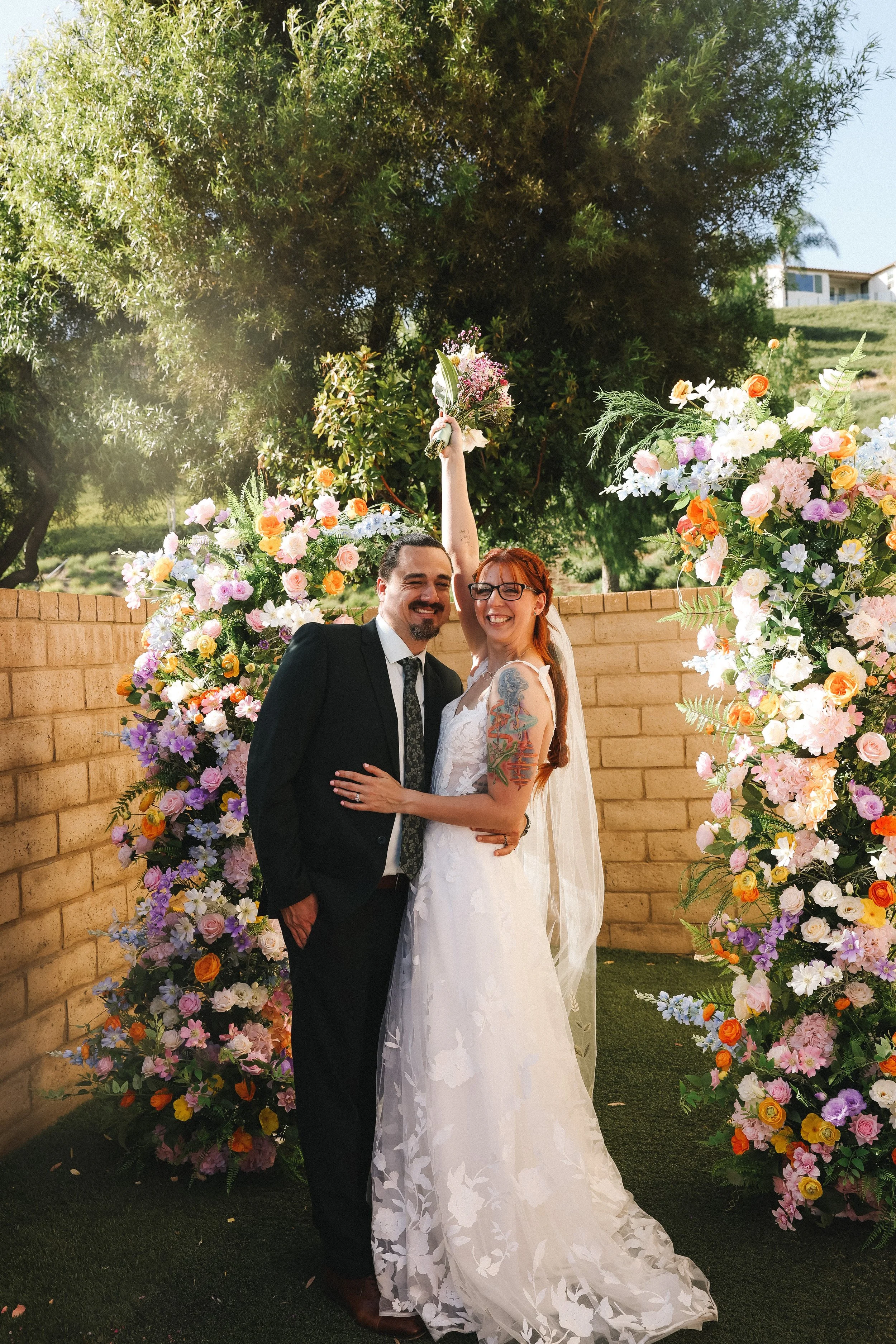 A floral archway made of colorful flowers such as roses, daisies, and other blossoms, set in a wooded outdoor area with trees and sunlight filtering through the branches.