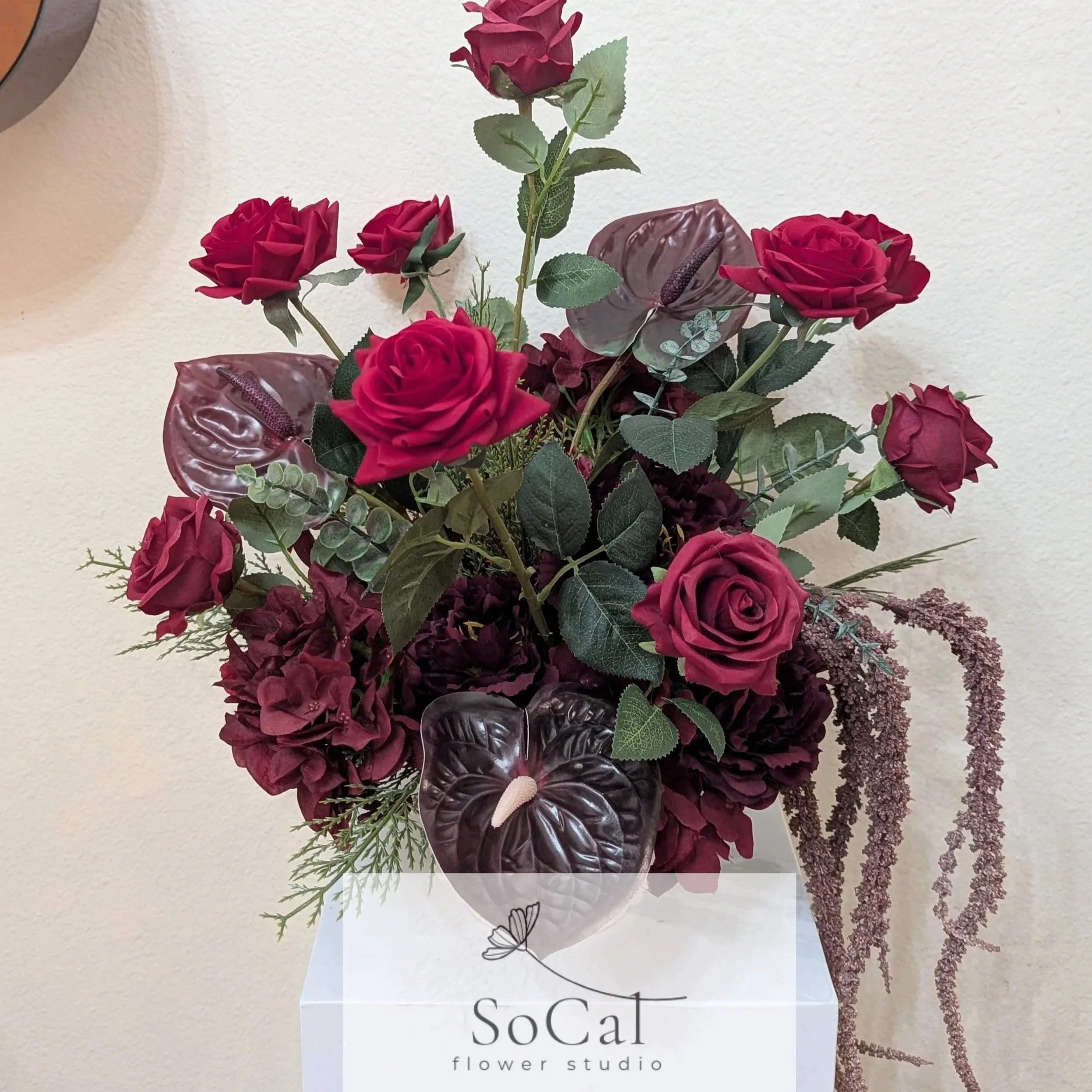 A floral arrangement with red roses, purple anthuriums, greenery, and hanging purple amaranthus in a clear glass vase, placed on a white display stand with 'SoCal flower studio' printed on it.