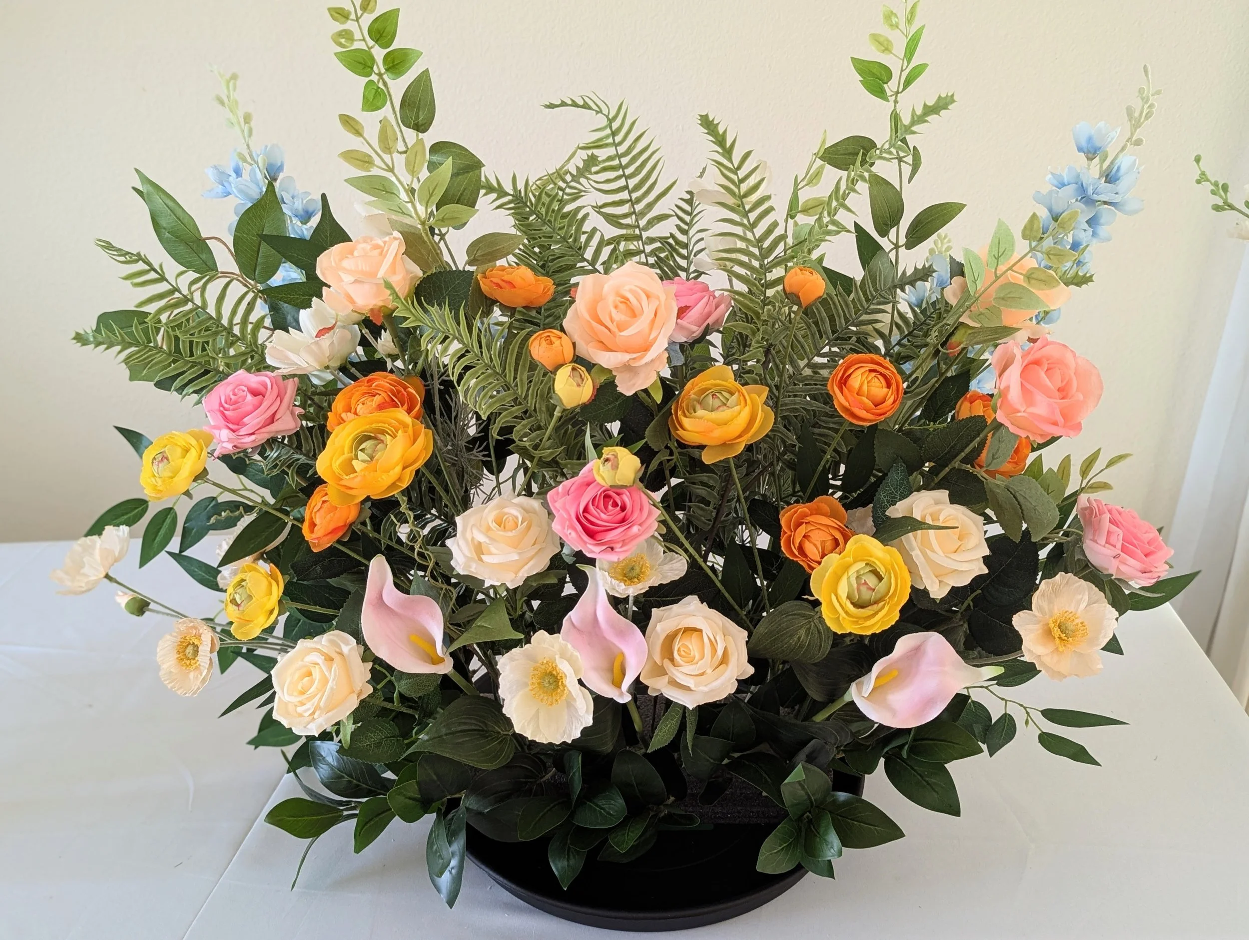 A large floral arrangement with pink, orange, yellow, white, and peach roses, calla lilies, and assorted greenery in a black container on a white surface.