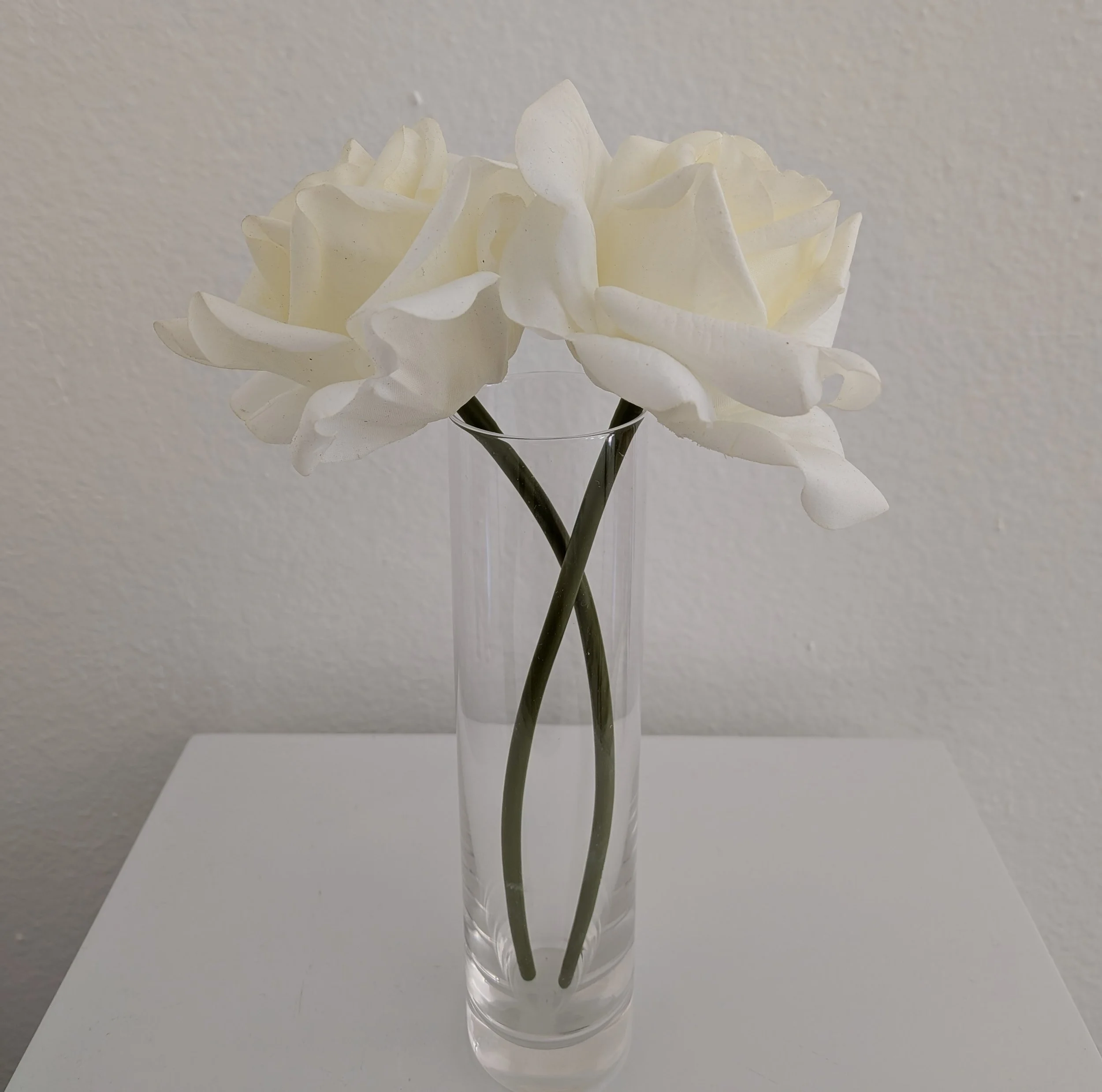 White artificial roses in a tall, clear glass vase on a white surface against a plain beige wall.