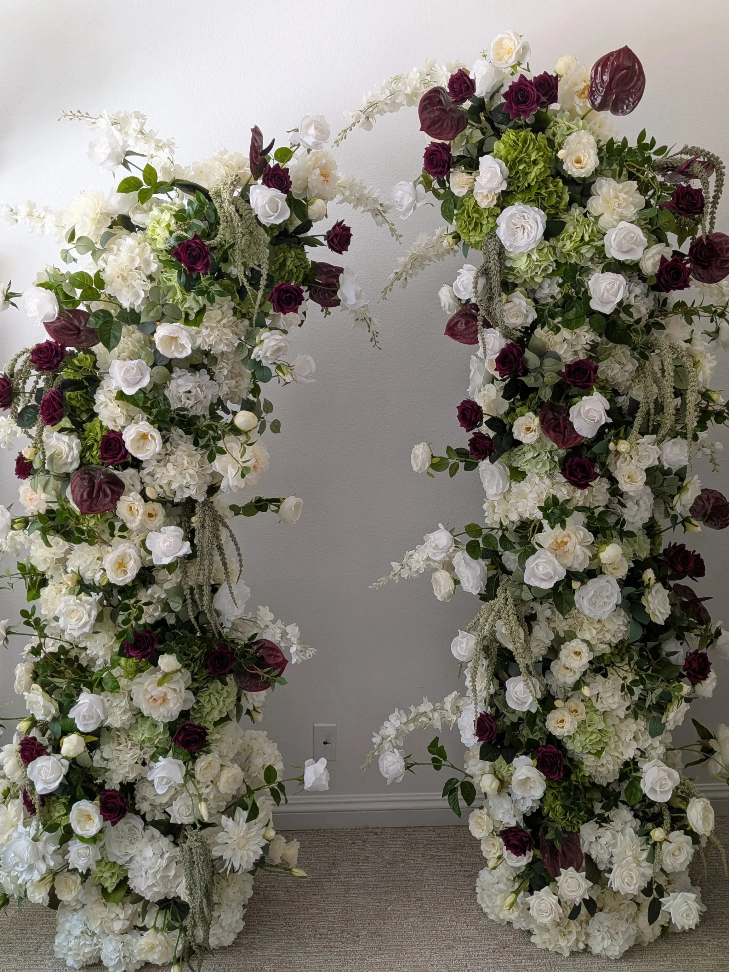 Large floral arrangement with white, burgundy, and light green flowers against a plain wall background.