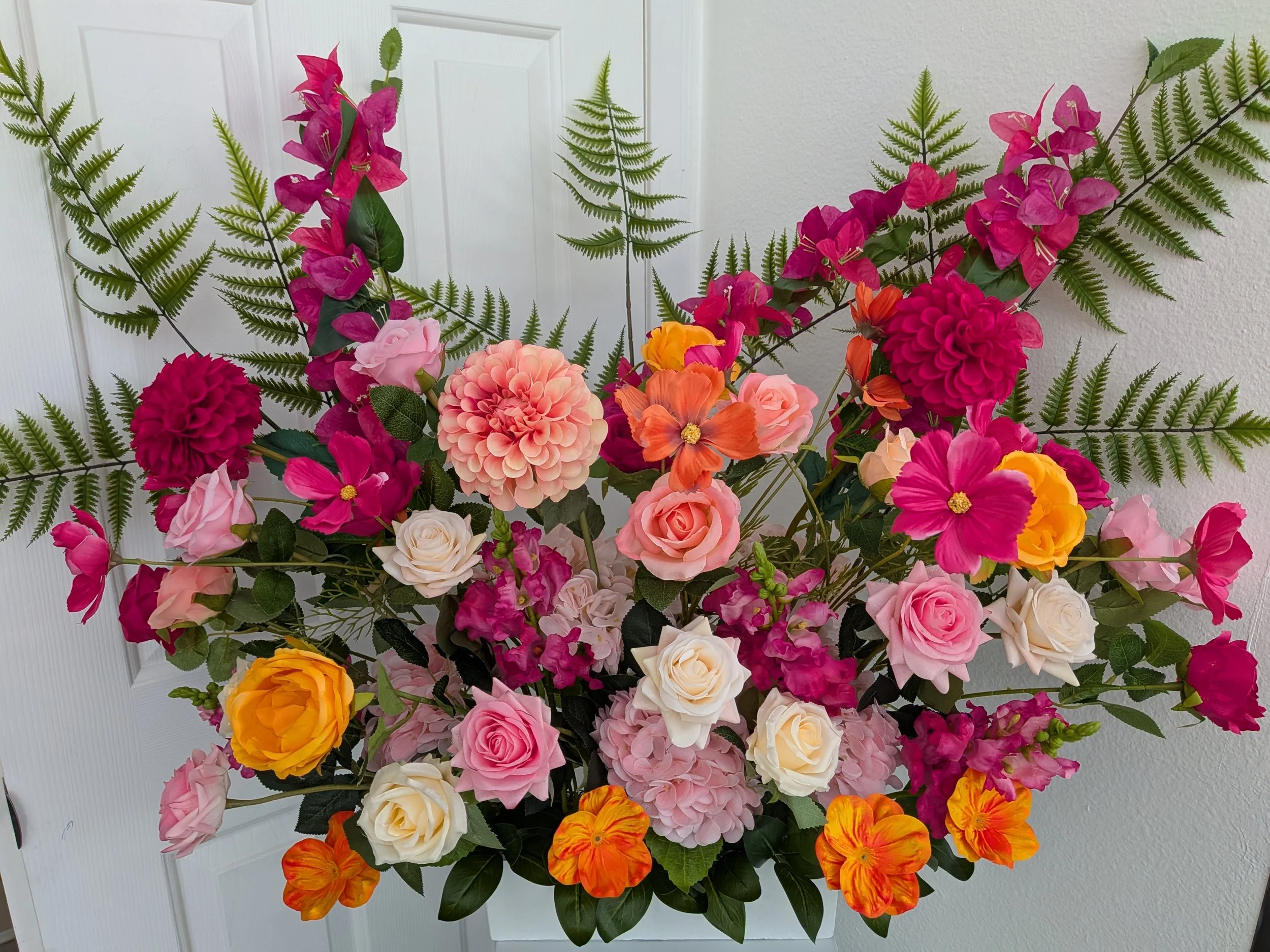 A large bouquet of artificial flowers including pink, red, orange, and white roses, pink bougainvilla, and orange and yellow pansies, with green fern fronds, arranged against a white door and wall background.