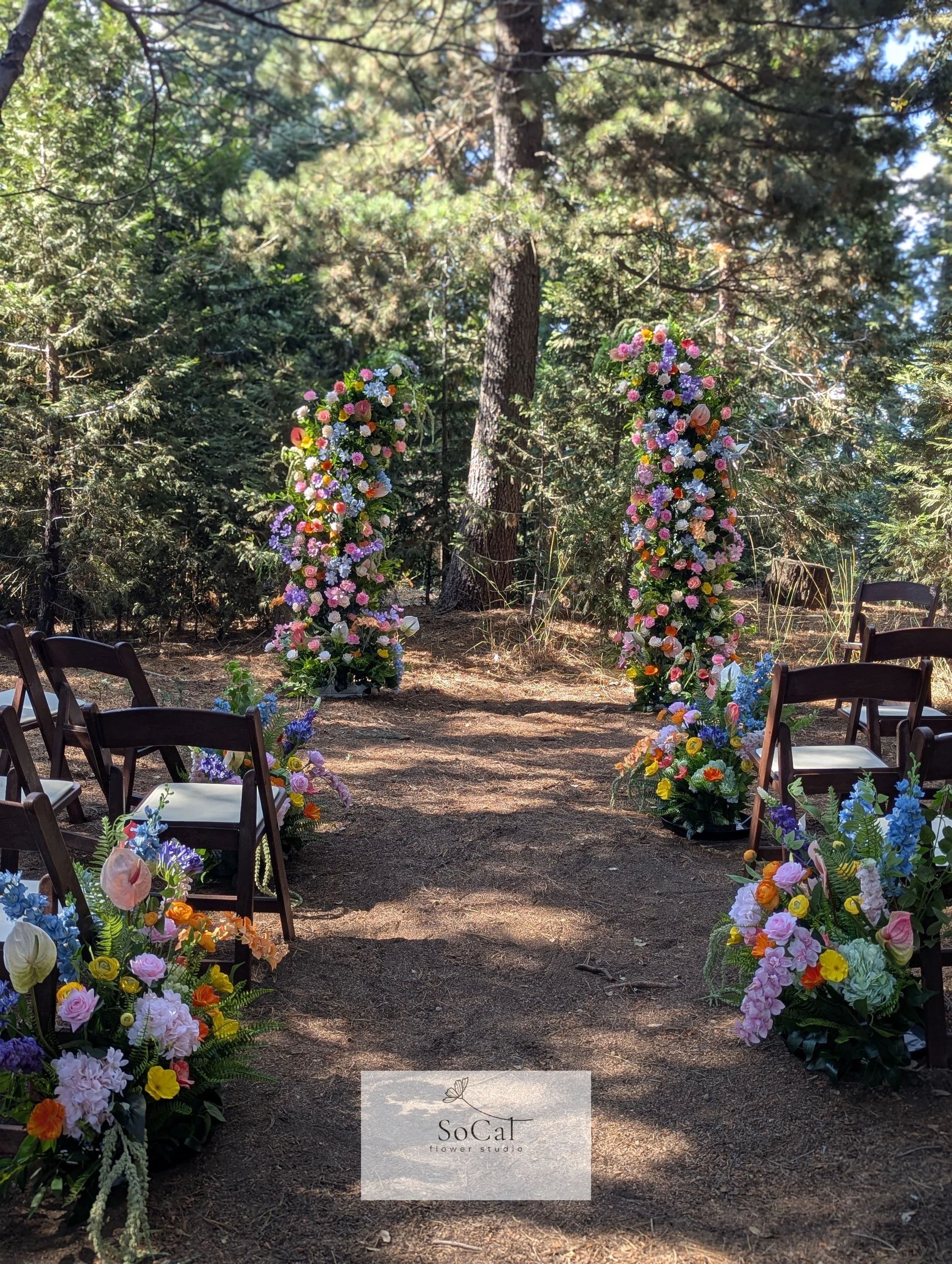 A floral archway made of colorful flowers such as roses, daisies, and other blossoms, set in a wooded outdoor area with trees and sunlight filtering through the branches.