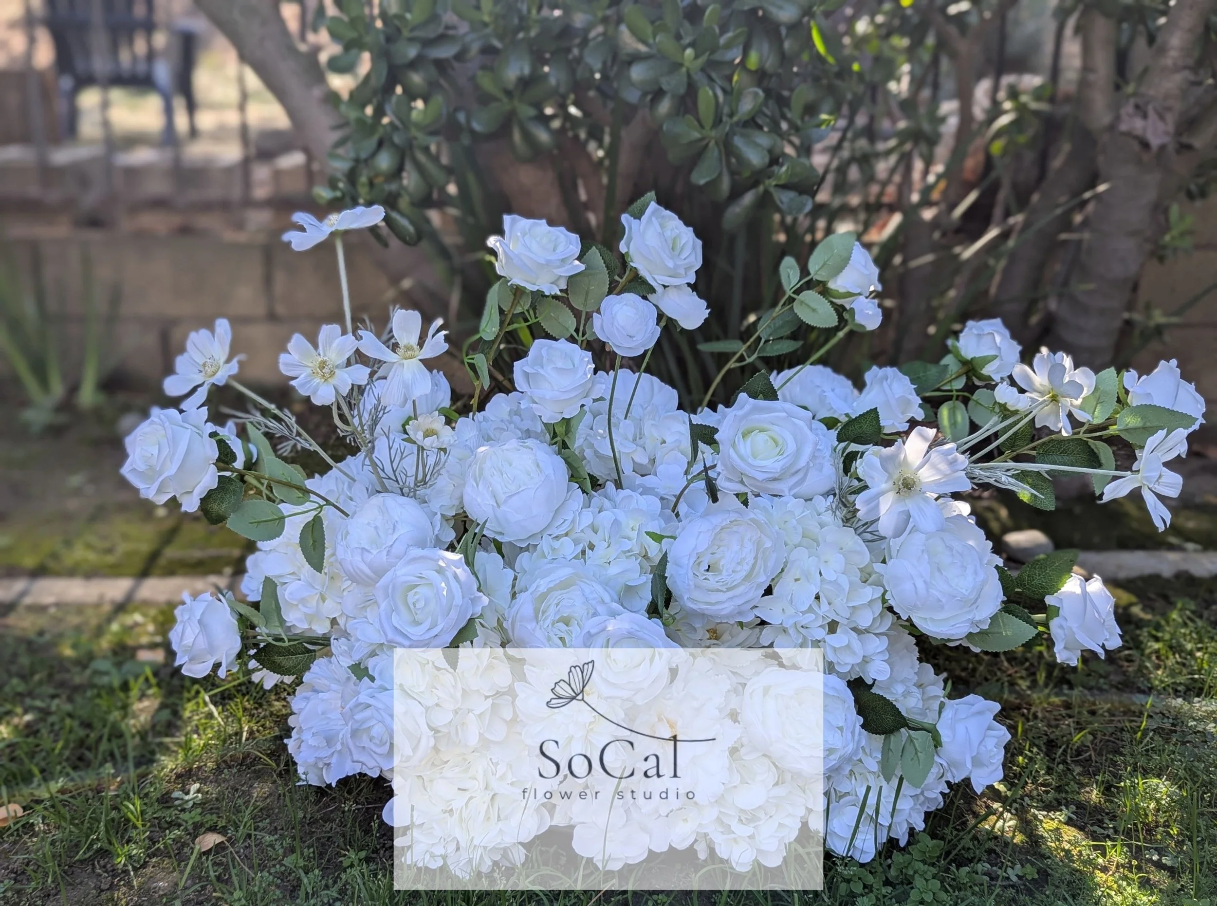 White artificial flower arrangement in a garden with a brick wall and plants in the background, with a SoCal Flower Studio logo overlay.