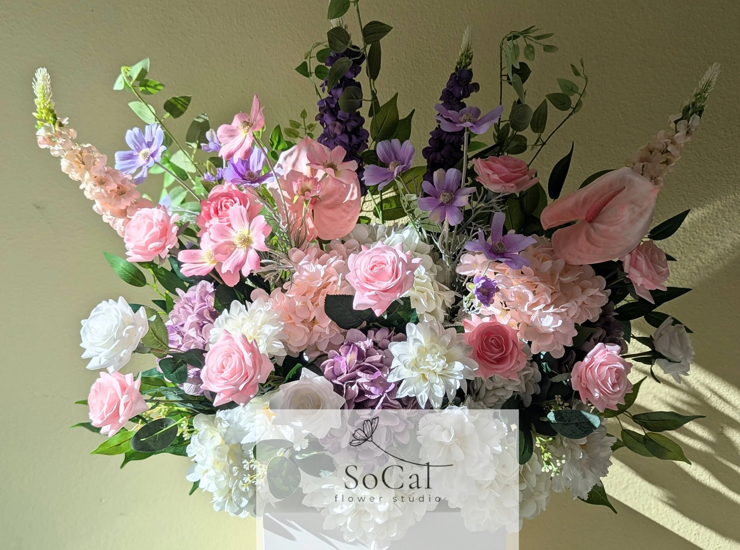 A bouquet of pink, purple, white, and cream flowers with green leaves, arranged in a vase with a "SoCal flower studio" label.