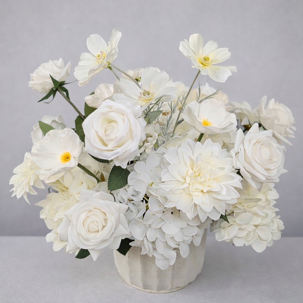A bouquet of white flowers including roses, poppies, dahlias, and other blooms in a white vase with a SoCal flower studio card, set against a light gray curtain background.