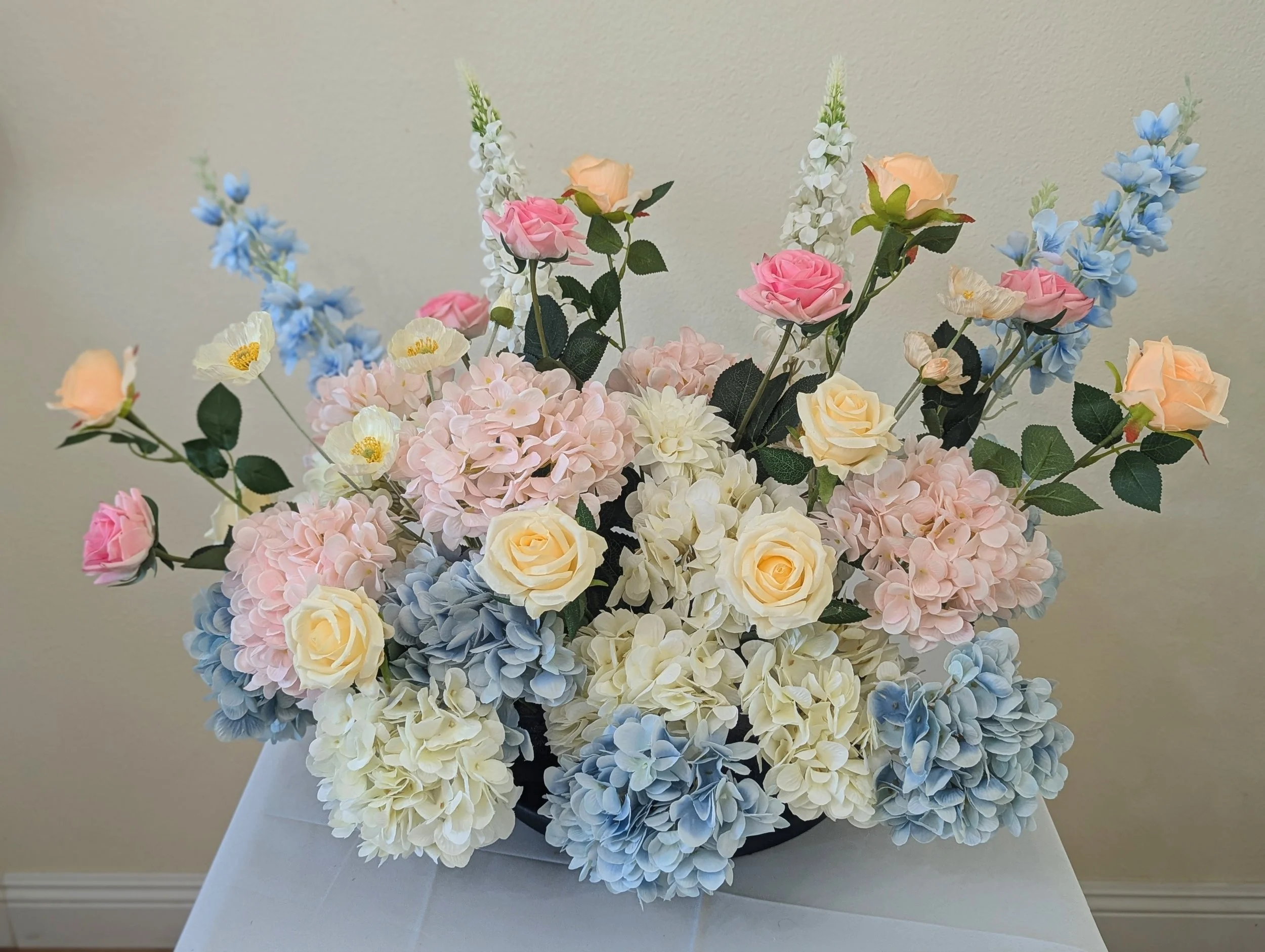 A large bouquet of pastel-colored flowers including roses, hydrangeas, and delphiniums in a white vase on a white table.