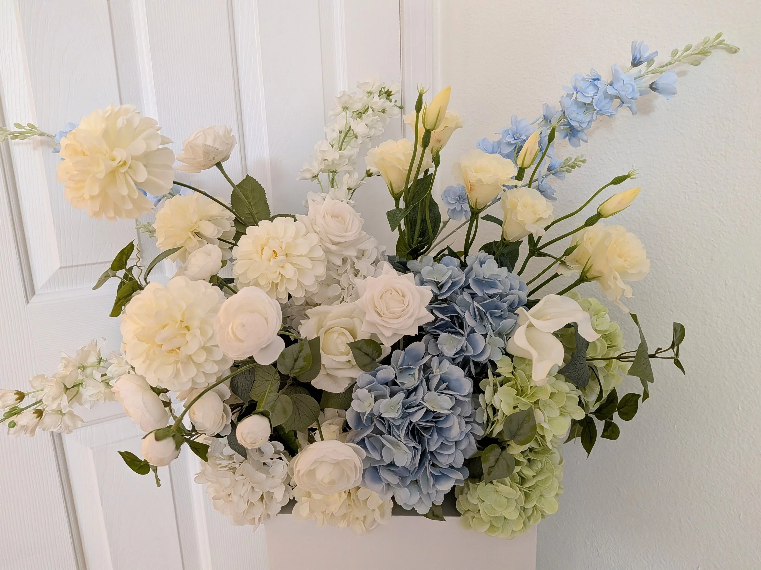 A bouquet of white, blue, and pale green flowers, including roses, hydrangeas, and delphiniums, set against a white background.