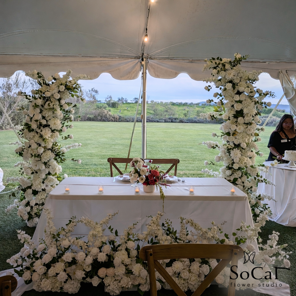 A floral arch decorated with white roses and greenery, set outdoors on a dirt path with trees and blue sky in the background.