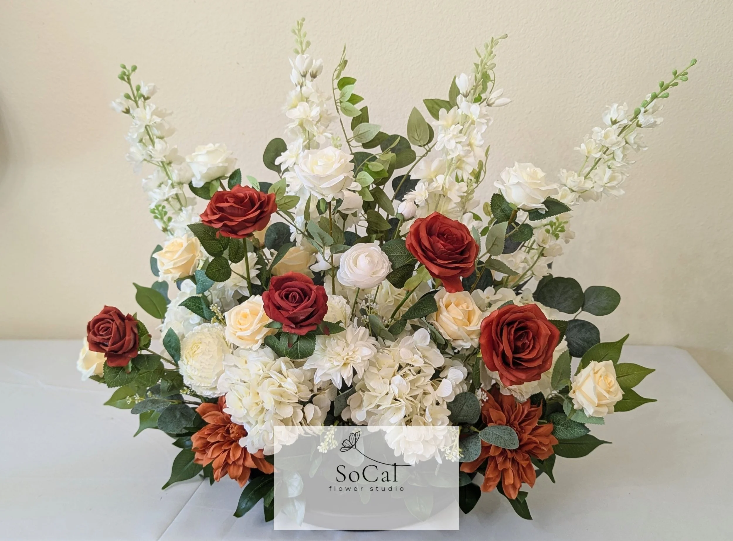 A floral arrangement with white, red, and orange flowers and green leaves in a white vase, with a "SoCal flower studio" logo at the bottom.