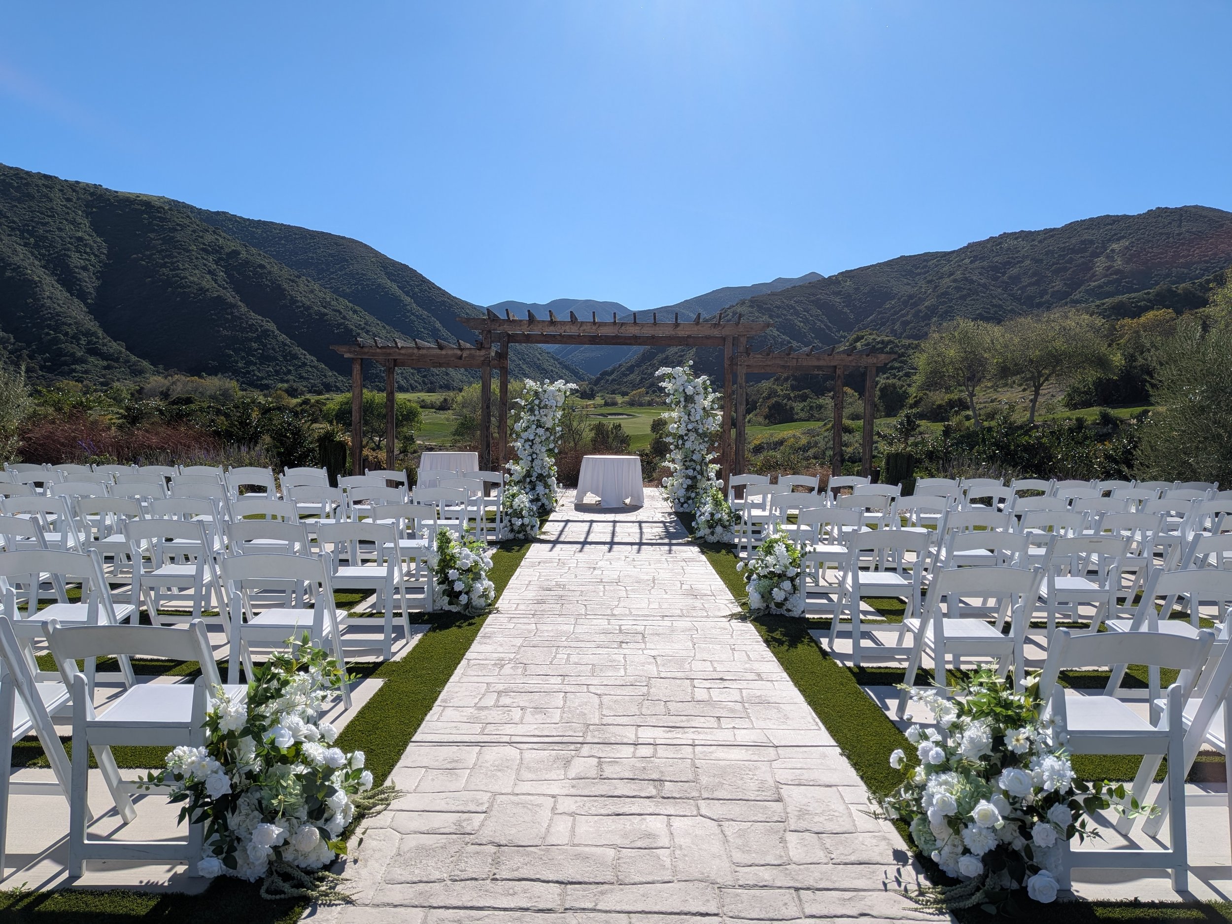 Outdoor wedding setup with white chairs, floral arrangements, and a wooden arch in a scenic mountainous area