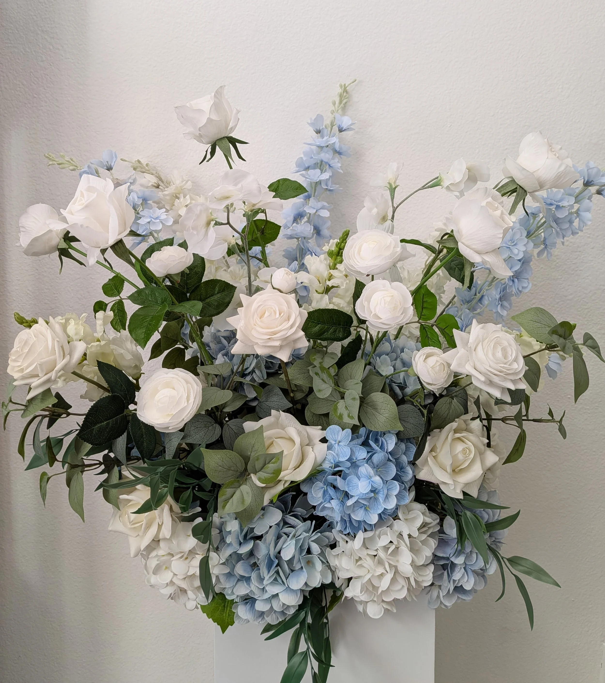 A large bouquet of artificial white roses, light blue hydrangeas, and greenery, arranged in a decorative display indoors.