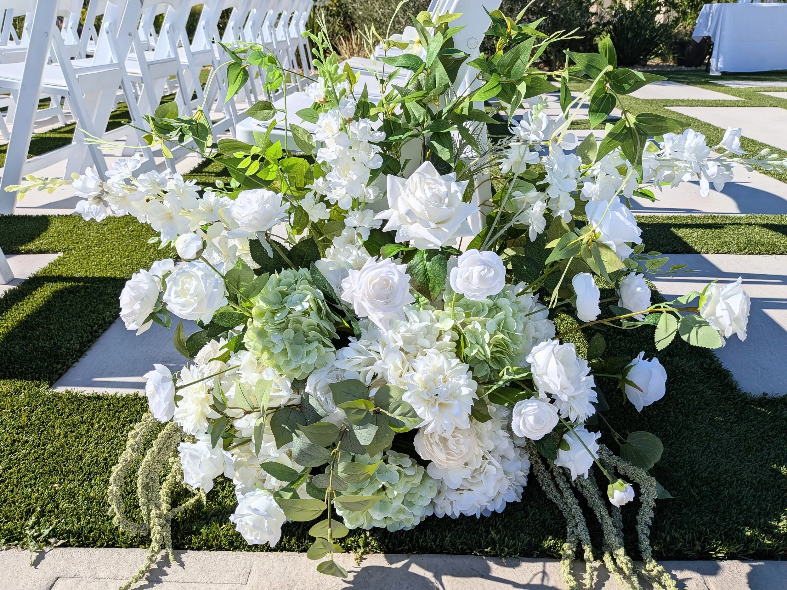 White floral arrangement with roses, hydrangeas, and greenery at an outdoor event.