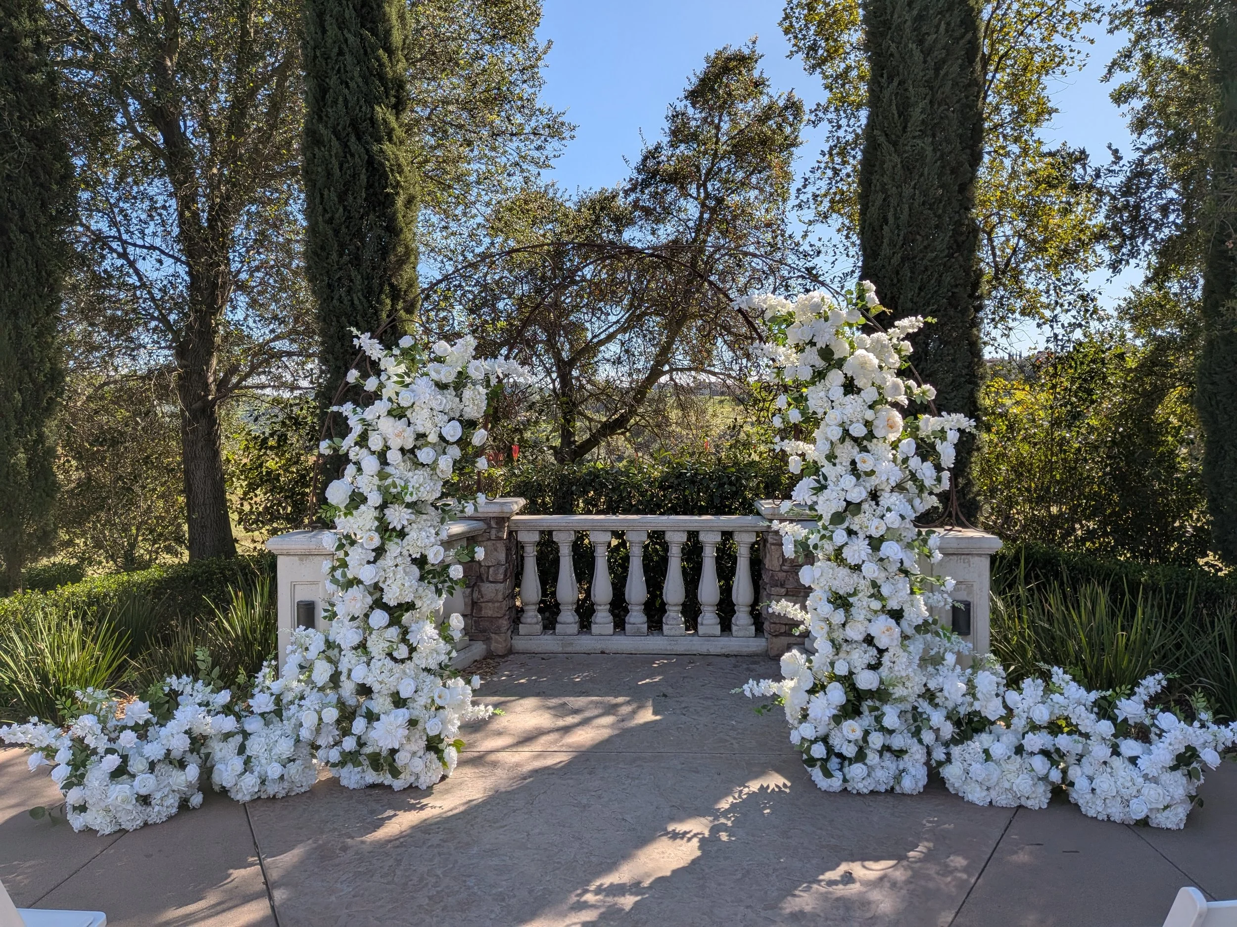 White floral wedding arch decorated with white roses and greenery, set on a stone platform with a stone and white balustrade, surrounded by tall trees and bushes in a garden.
