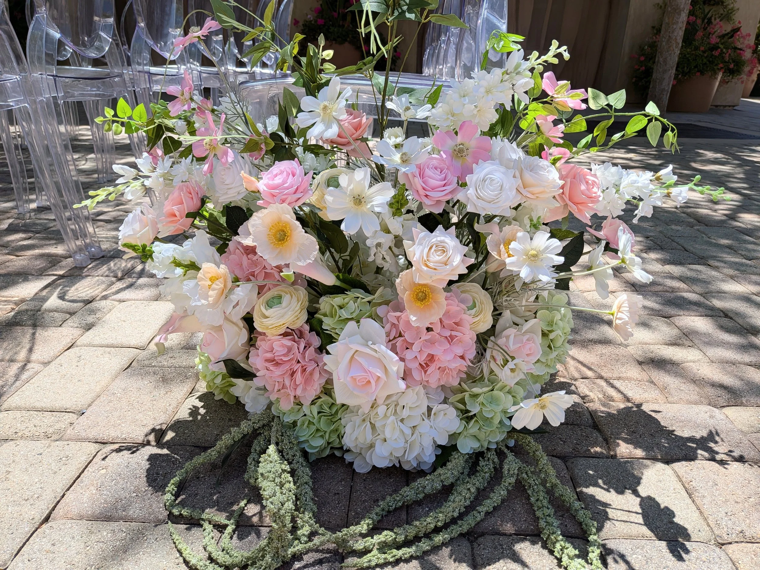 A floral arrangement with pink, white, and cream roses, hydrangeas, and other flowers, placed outdoors on a paved surface.