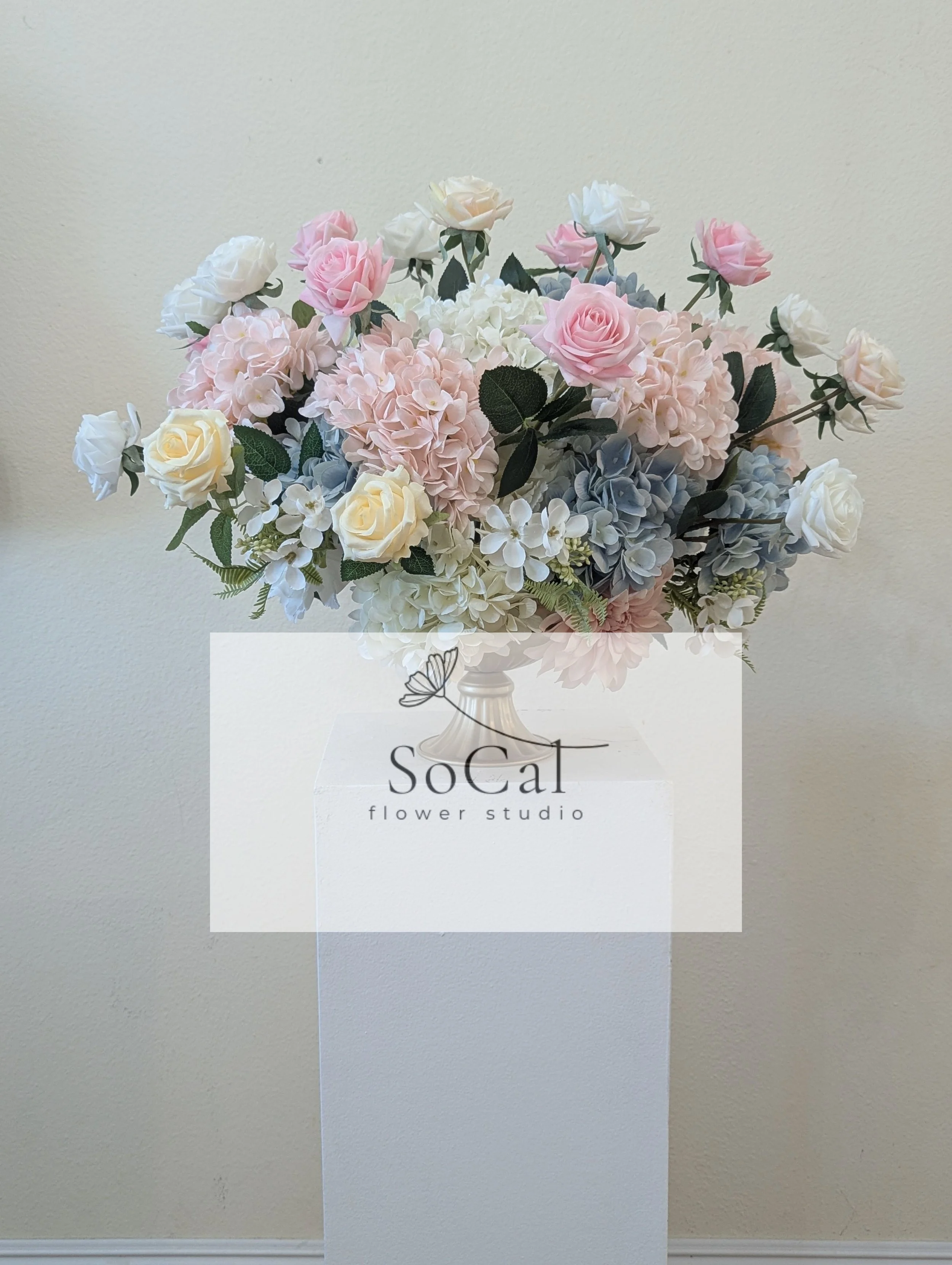 A large bouquet of pastel pink, white, blue, and cream roses, hydrangeas, and other flowers in a white vase, with a SoCal flower studio logo overlay.