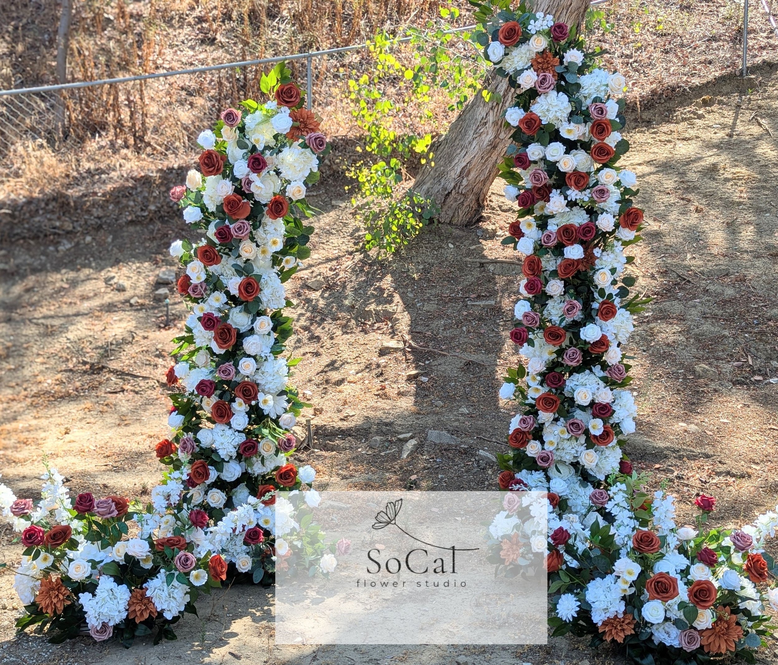 A large floral arch made of red, white, pink, and purple roses along with white and peach-colored flowers, outdoors on dirt ground with some greenery and a tree in the background. The photo is from SoCat flower studio.
