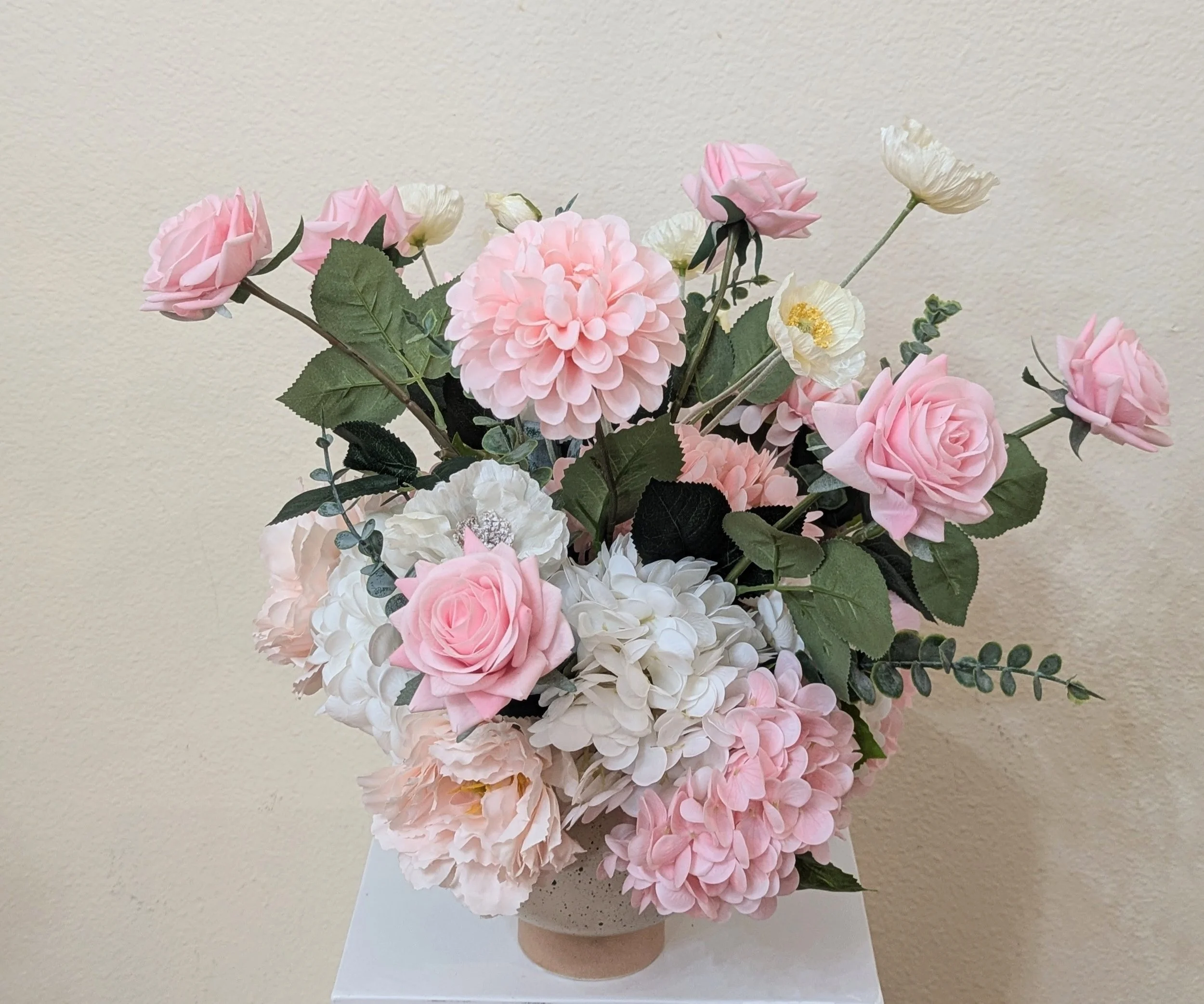 A bouquet of pink and white roses and hydrangeas in a white box with a 'SoCal flower studio' label, against a beige wall.