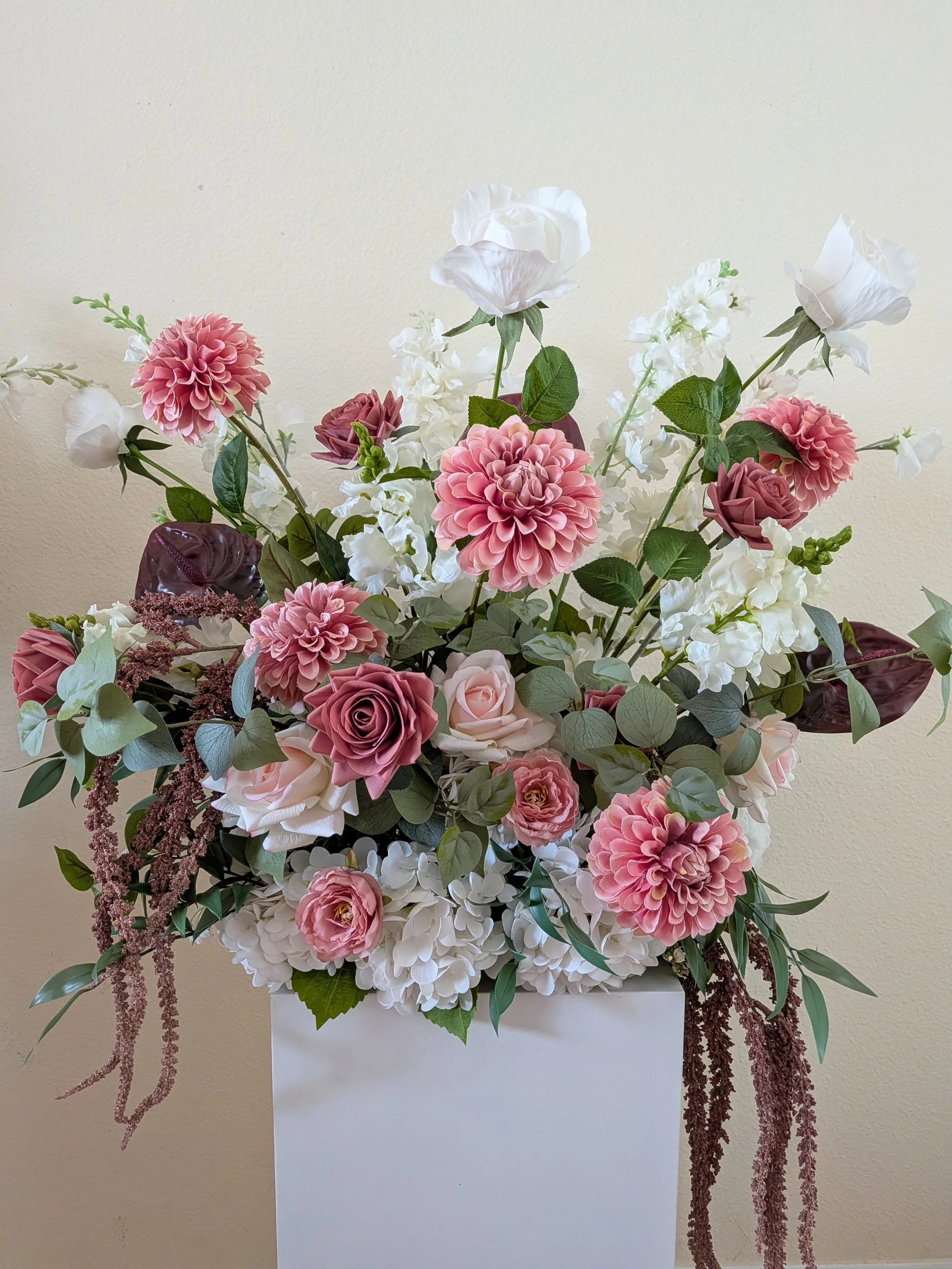 A floral arrangement with pink, white, and purple flowers and green foliage in a white container against a beige wall.