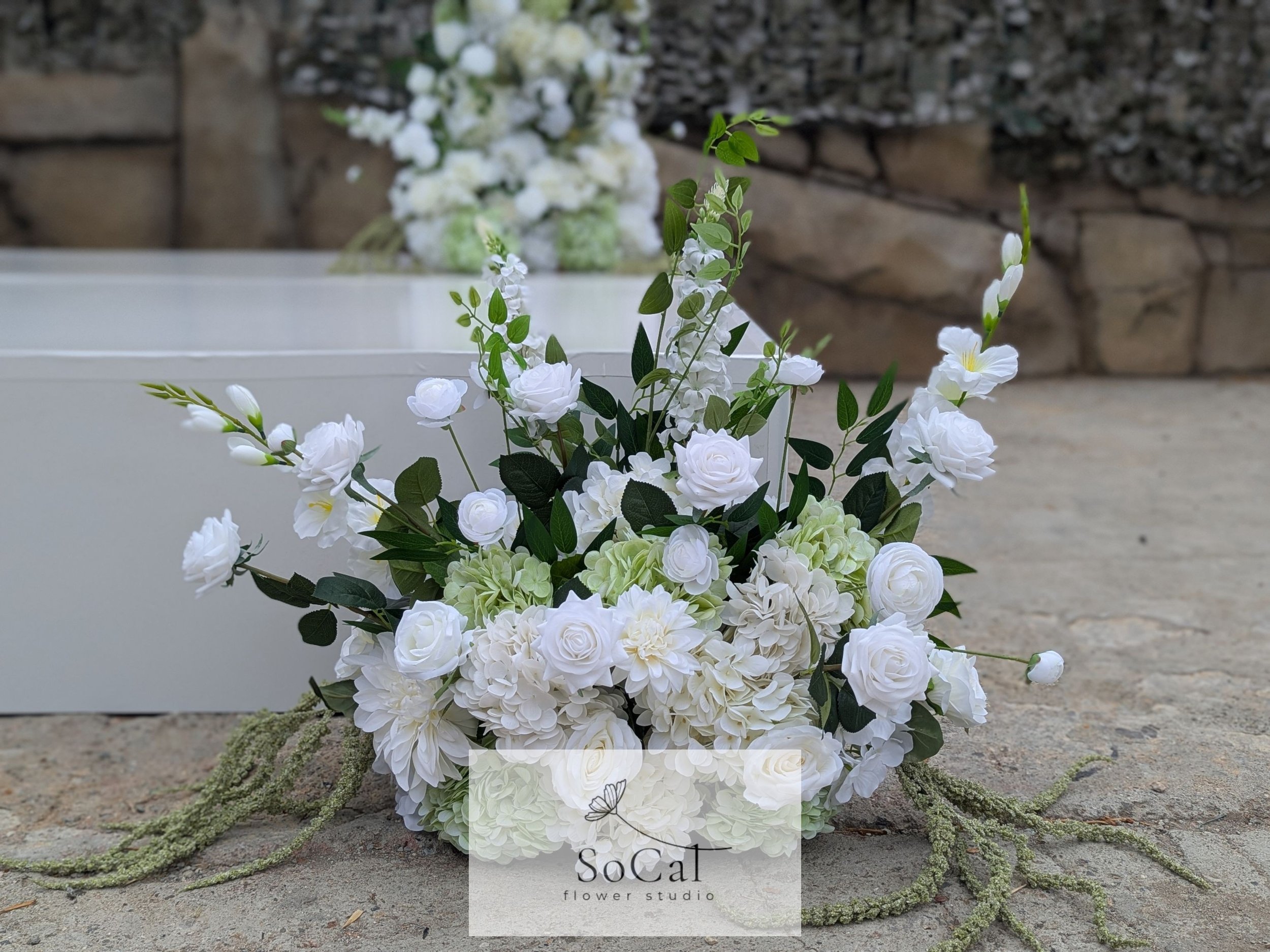 A white floral arrangement with roses, hydrangeas, and greenery, placed on a concrete surface in front of a blurred background with a stone wall and white boxes.