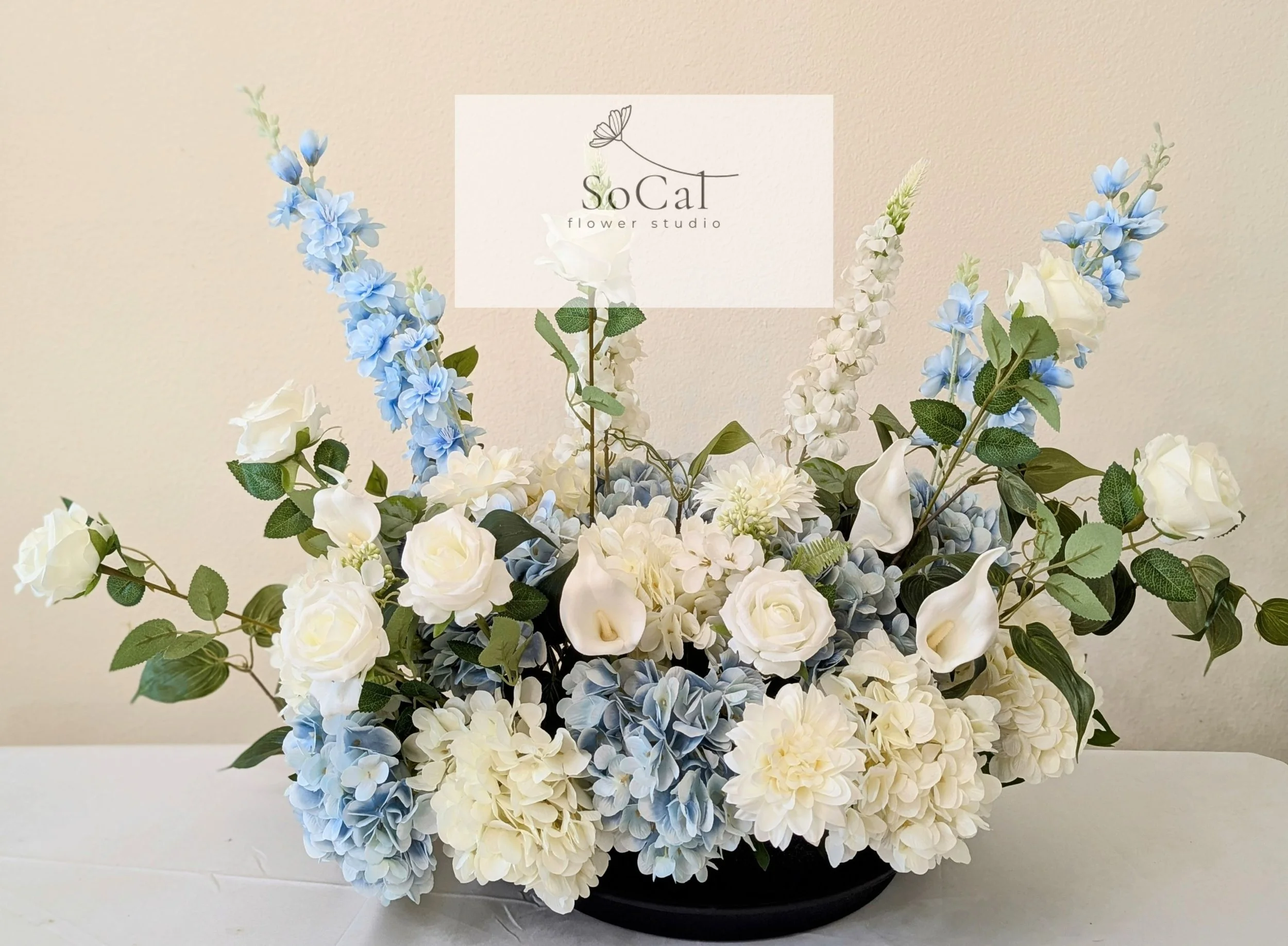 Arrangement of white roses, blue hydrangeas, and white calla lilies with green leaves, with a sign that reads "SoCal flower studio."
