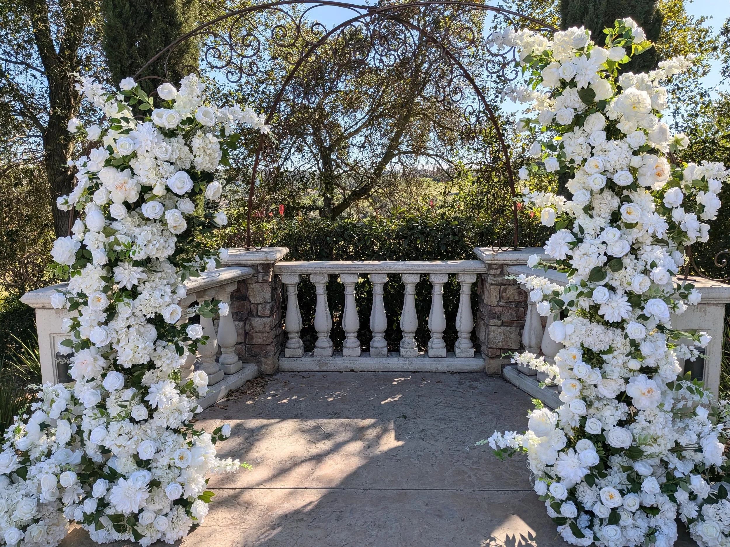 A small stone and concrete outdoor balcony or patio area decorated with white flowers on a metal arch and surrounding the railing, with trees and bushes visible in the background.