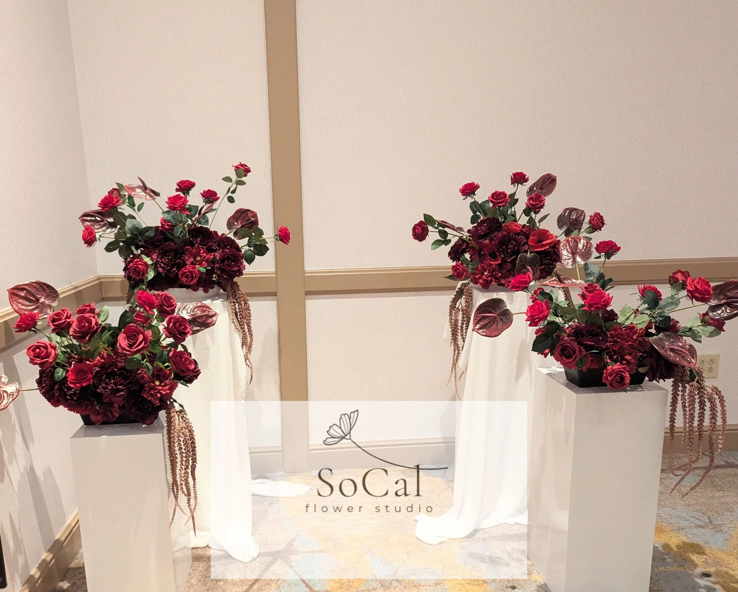 Three tall white planters with lush red and burgundy flowers, arranged on white fabric-covered pedestals in a room with beige walls and beige and gray patterned carpet. A transparent overlay with the text 'SoCal flower studio' and a small butterfly g