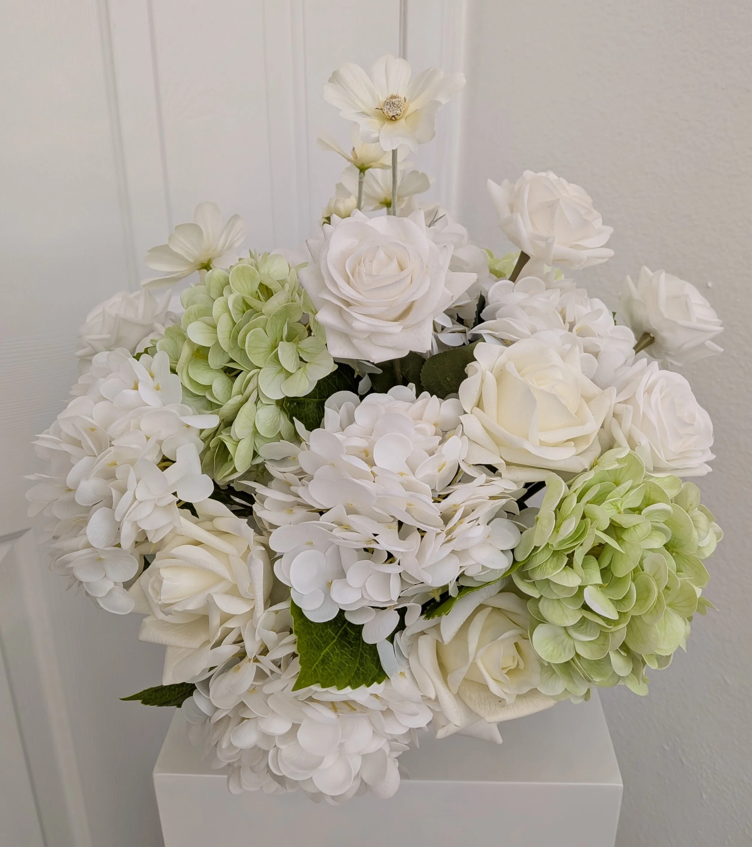 A white floral centerpiece featuring roses, hydrangeas, and greenery on a table in an event space.
