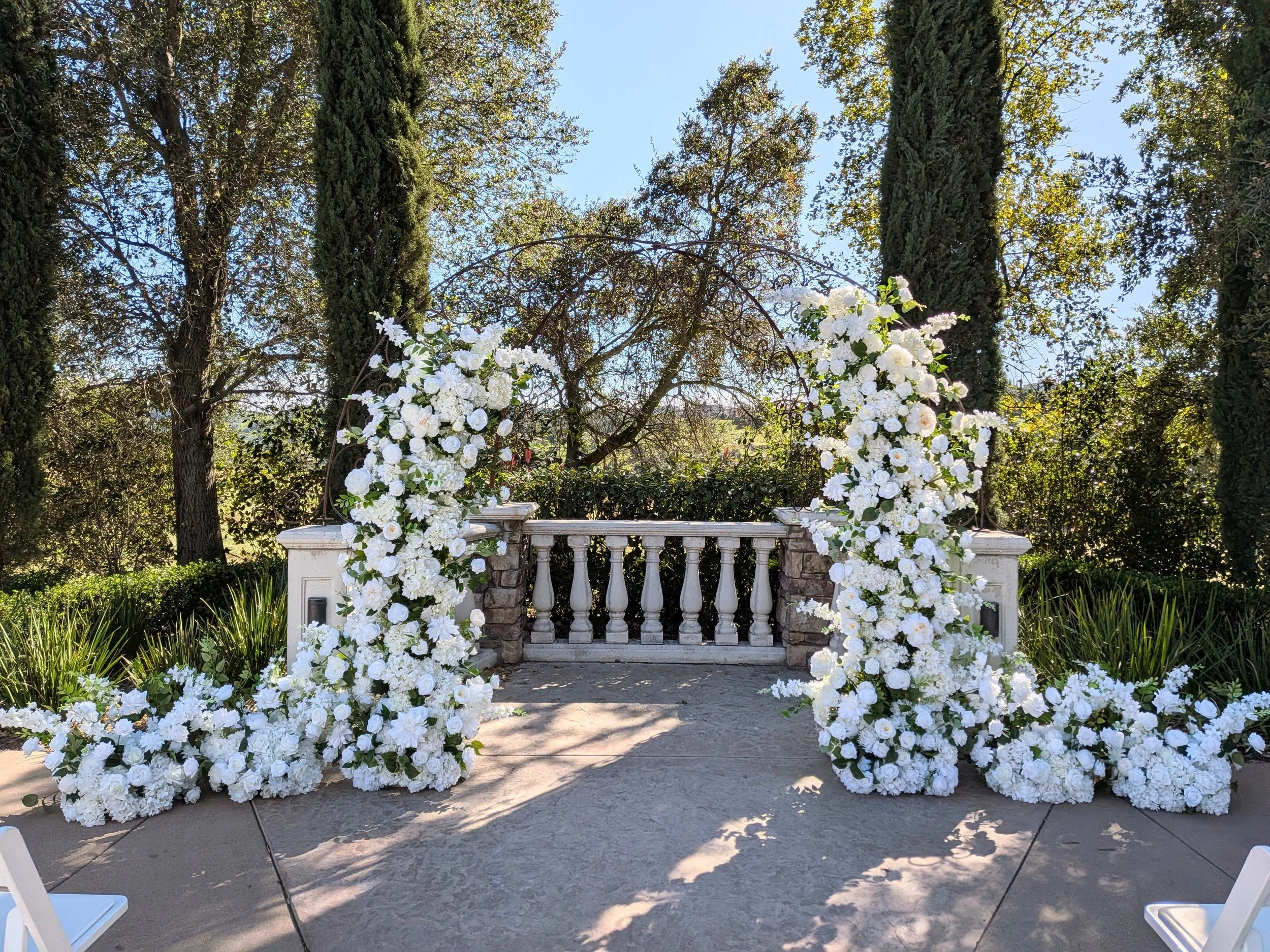 A floral arch decorated with white roses and greenery, set outdoors on a dirt path with trees and blue sky in the background.