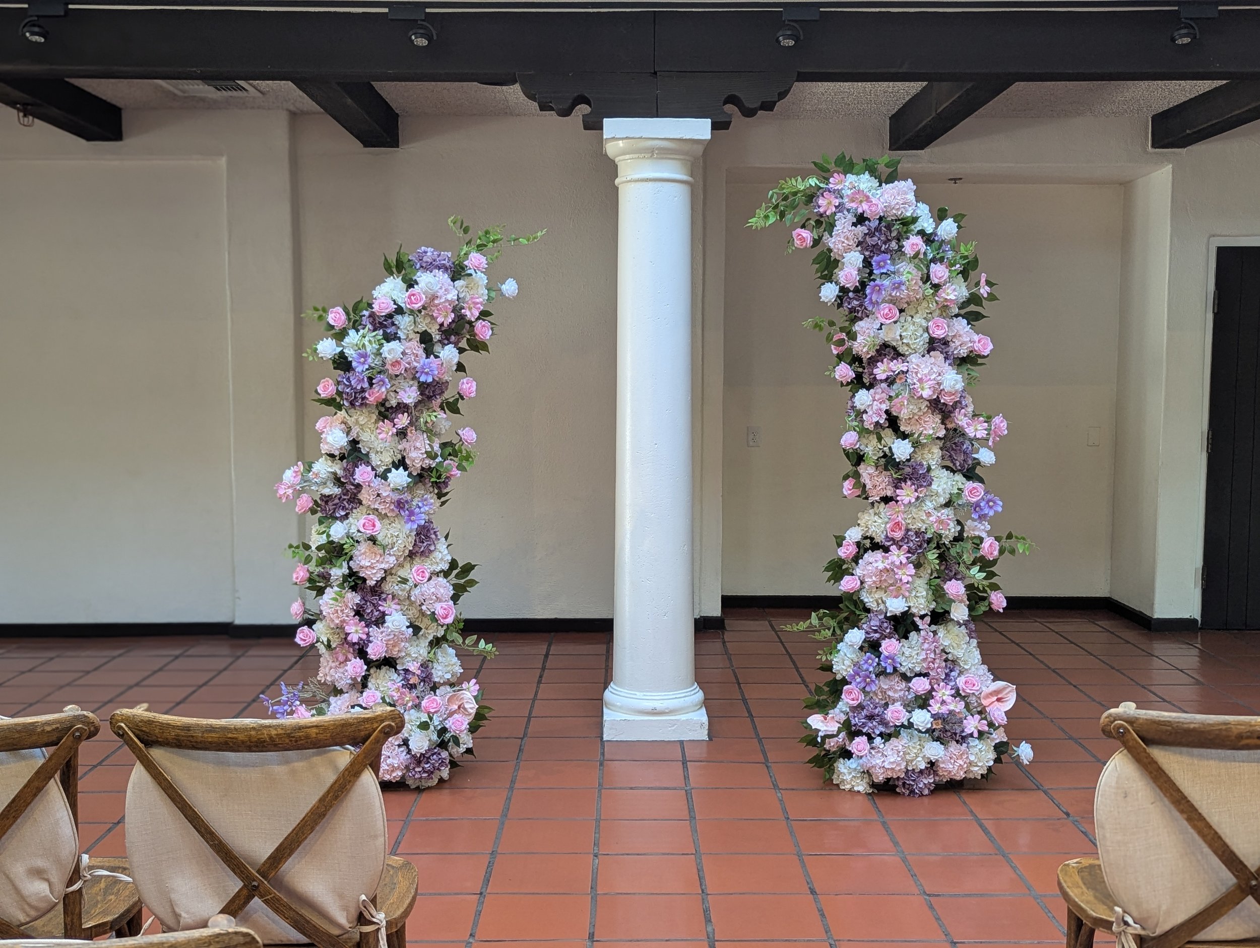 Two large floral arrangements with pink, purple, and white flowers on either side of a white column inside a room with terracotta tile flooring and beige walls.