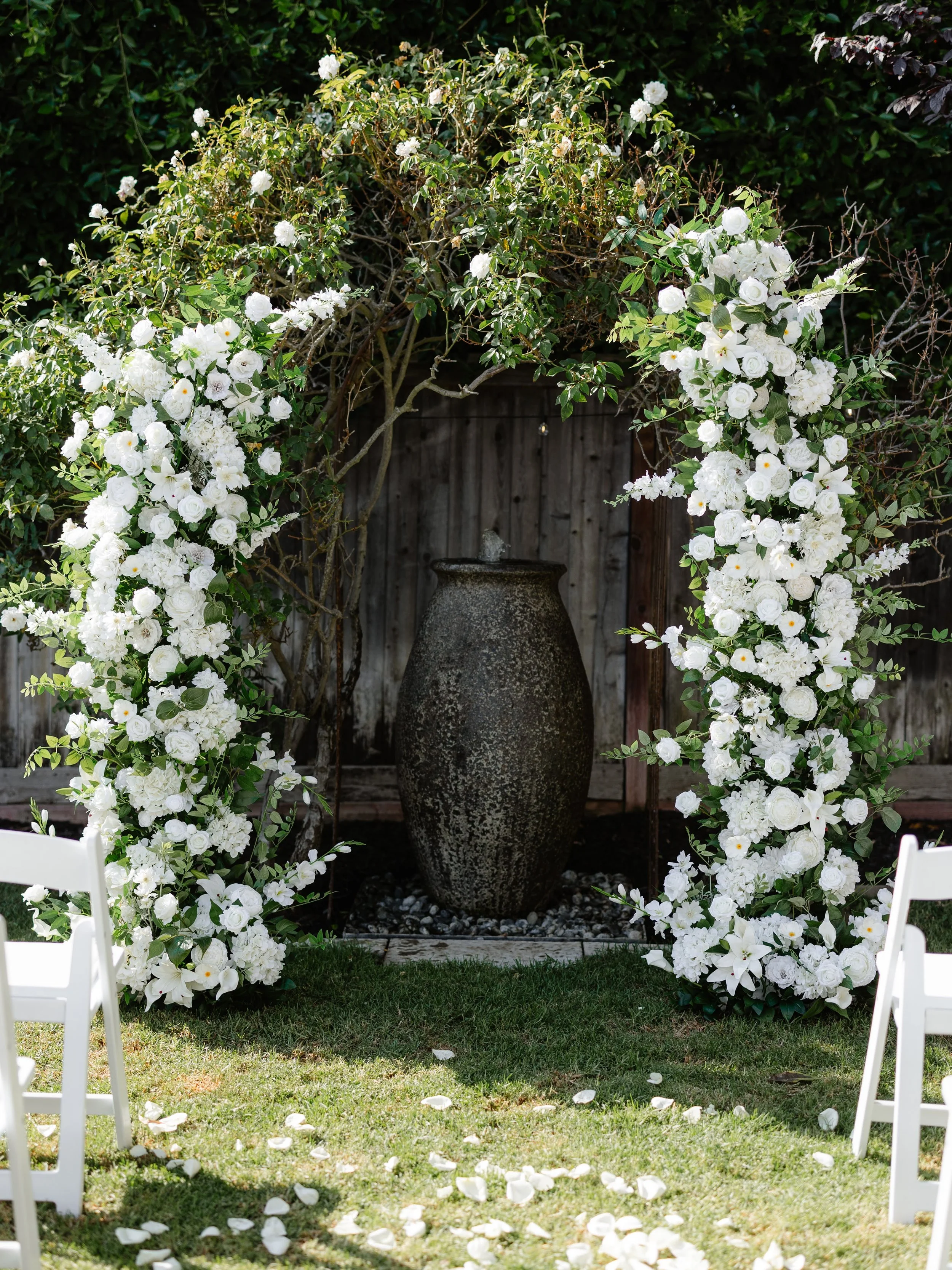 A wedding arch decorated with white flowers, surrounding a fountain on a grassy lawn.