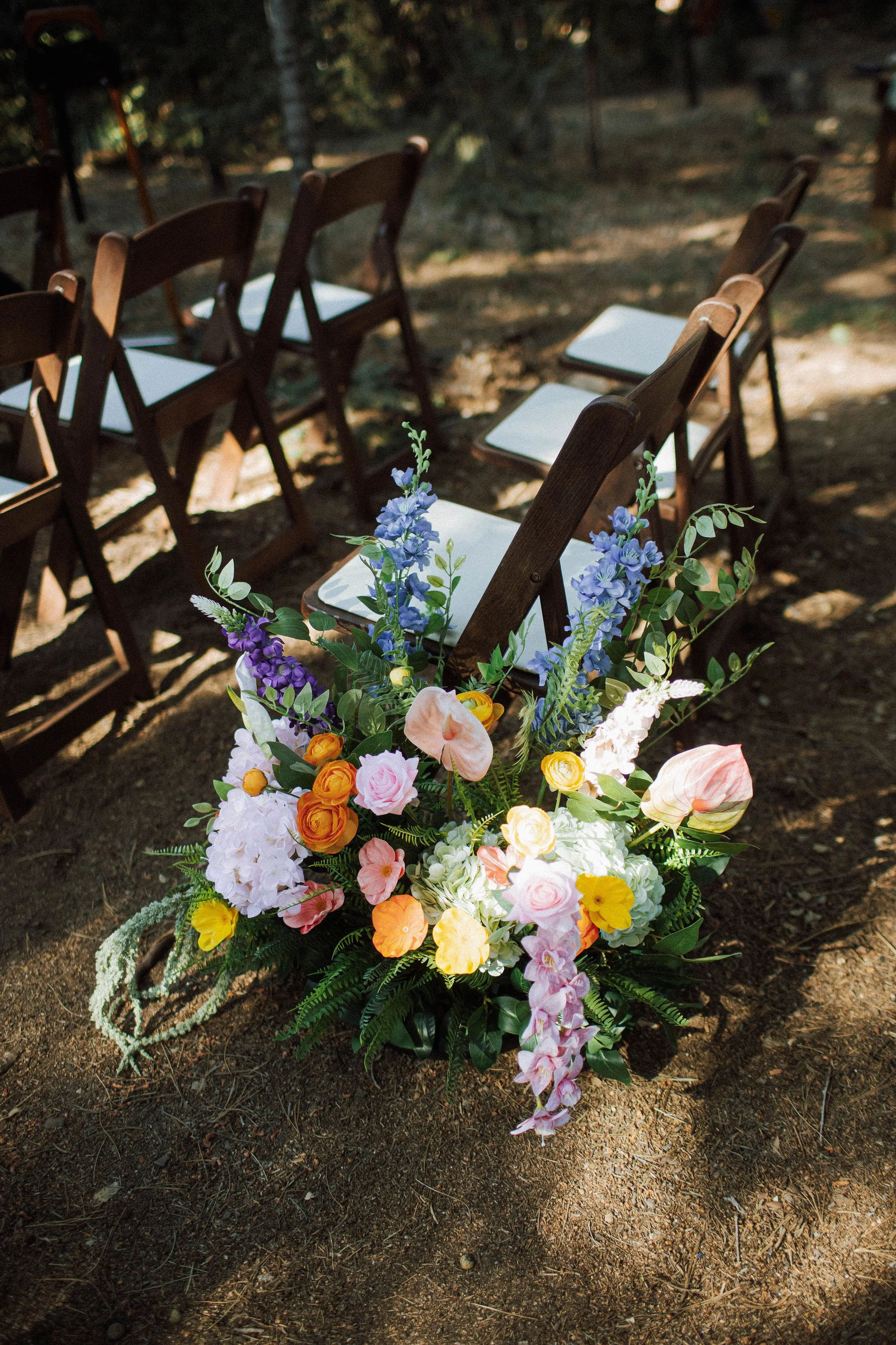 A colorful floral arrangement featuring pink roses, orange flowers, blue delphiniums, pink anthuriums, green hydrangeas, and greenery in a black vase on a wooden surface.