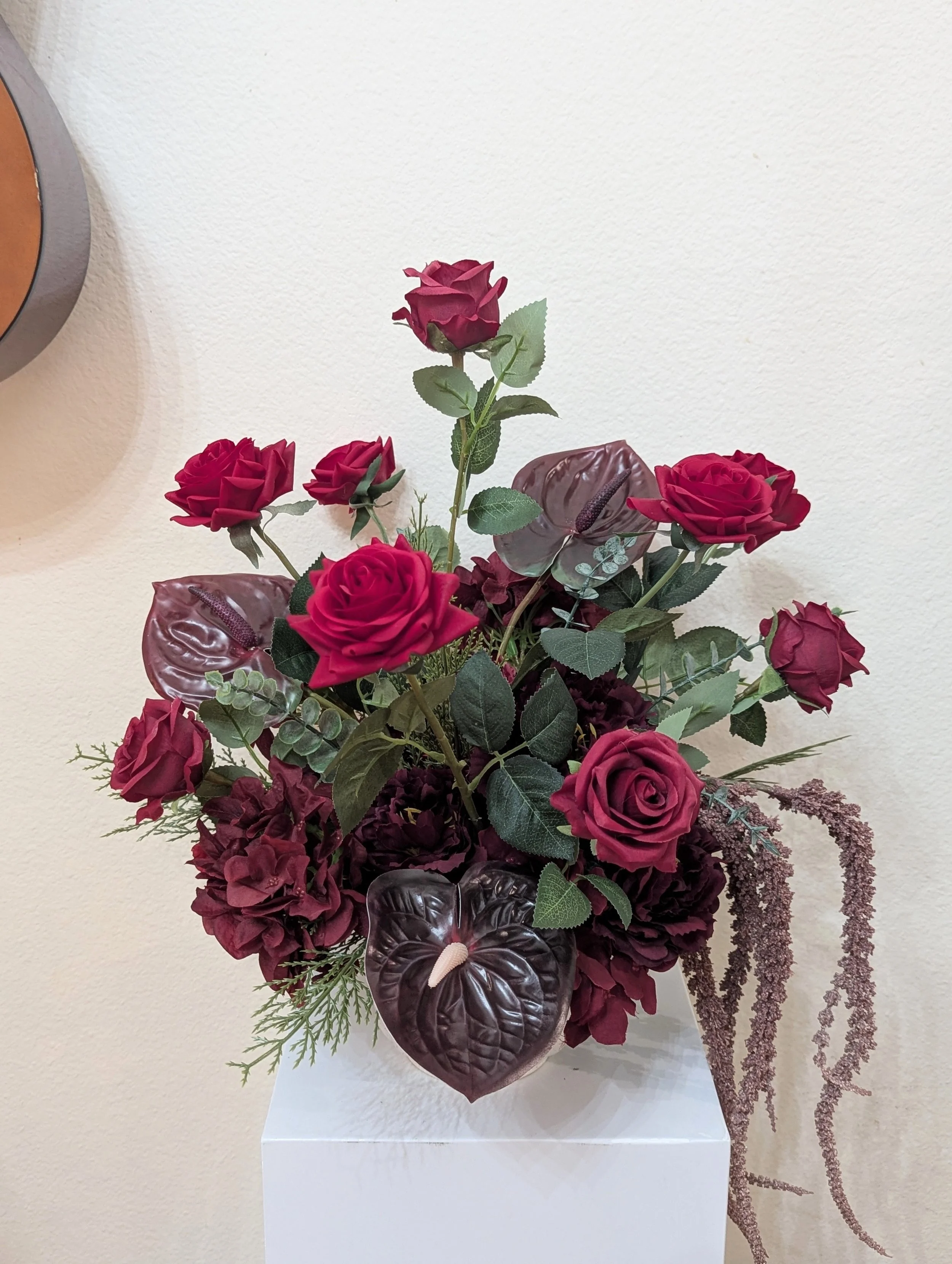 Arrangement of dark red hydrangeas, black calla lilies, and purple-leaved plants in a white square vase on a plain wall background.